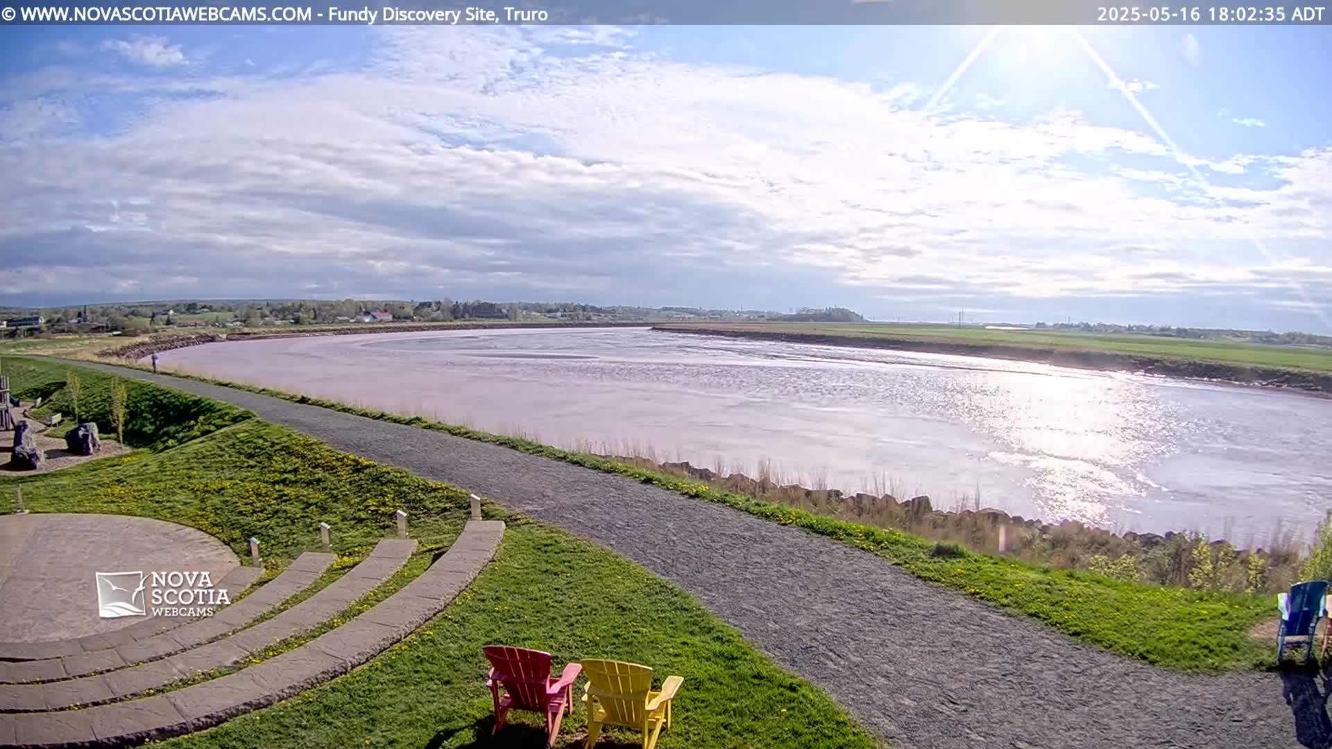 A partly cloudy day reveals a wide, calm river reflecting sunlight, alongside a paved path and grassy area with a few chairs.
