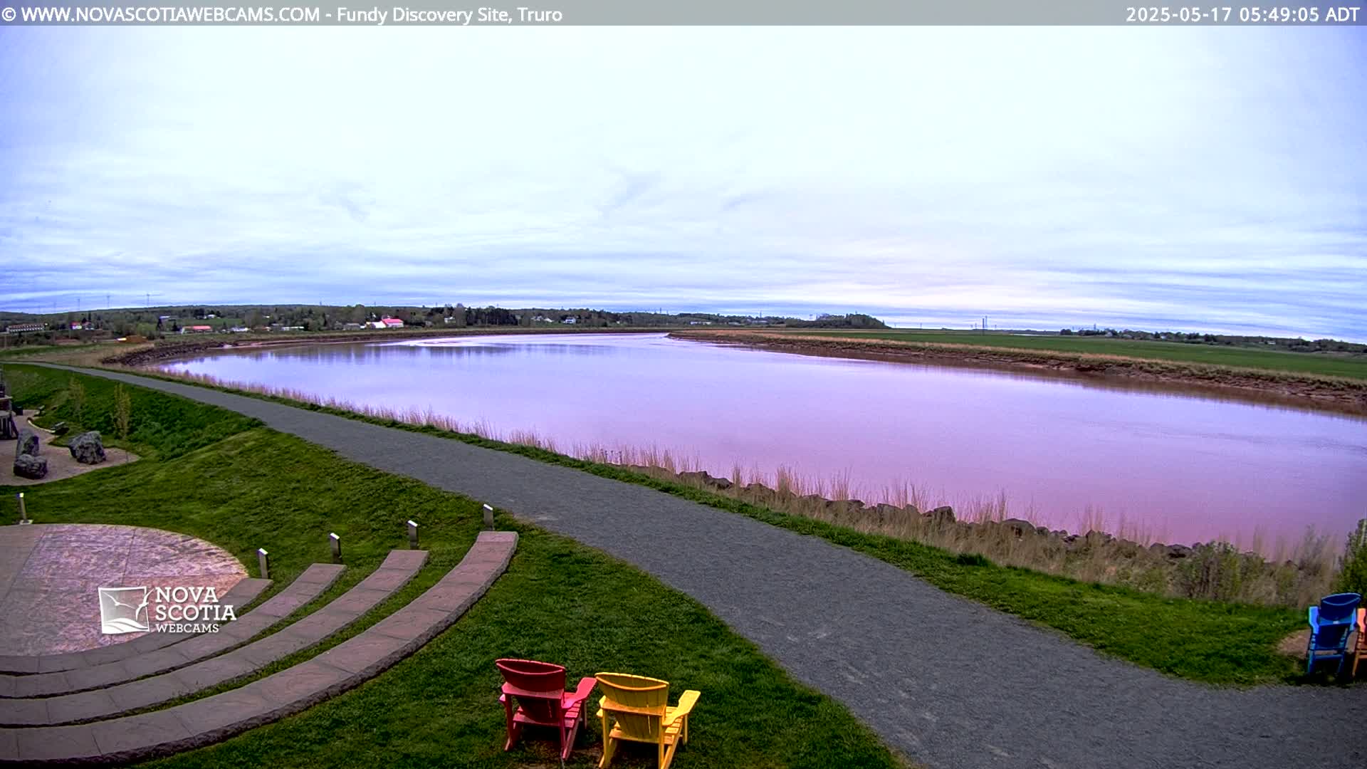 A paved path winds alongside a calm, pinkish-purple river, with green grassy areas and a few colorful chairs in the foreground under a mostly clear, light-blue sky.