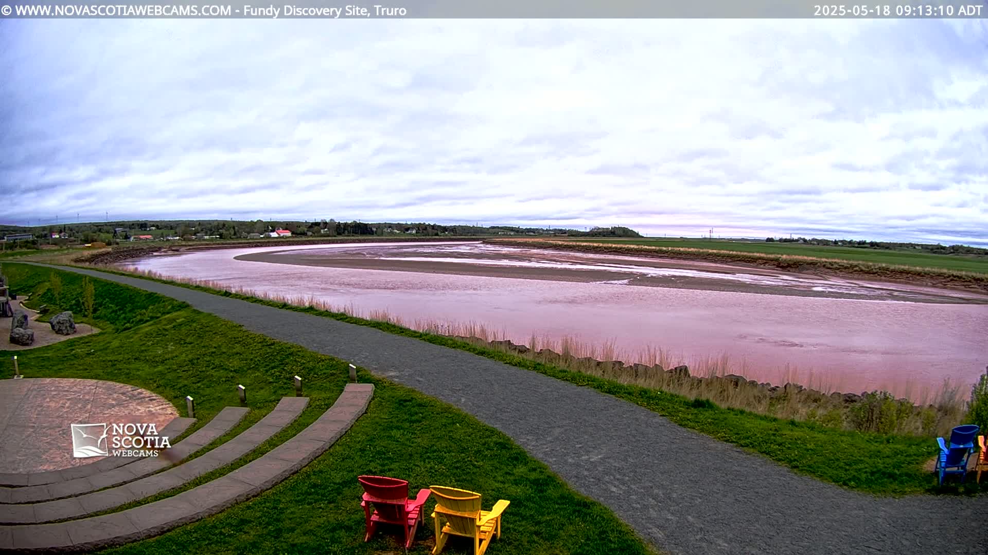 A pinkish-red river winds through a grassy landscape under a partly cloudy sky, with a paved pathway and several colorful Adirondack chairs in the foreground.
