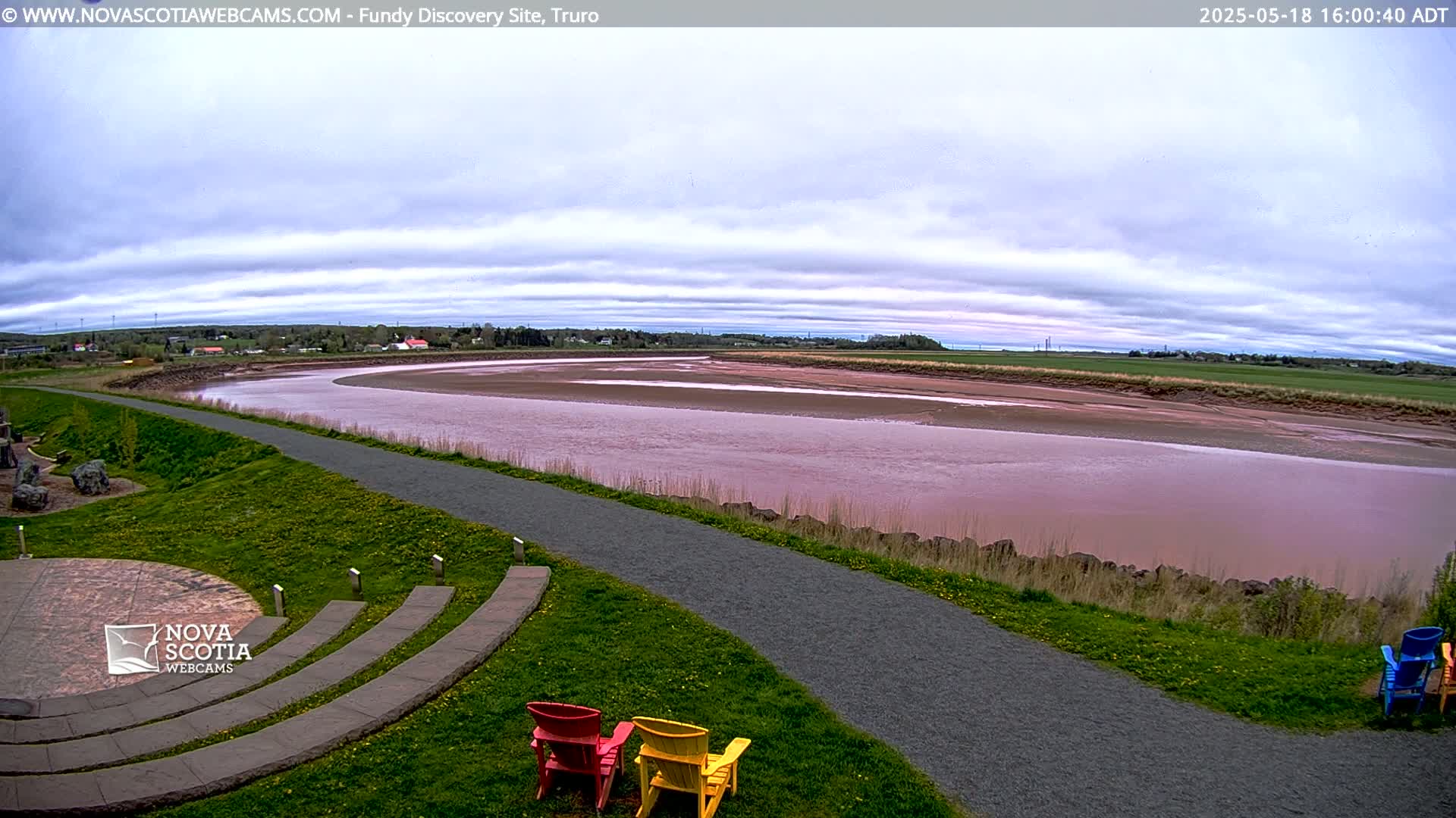 A wide, pinkish-red river curves gently under a mostly cloudy sky, with a paved path and several colorful Adirondack chairs situated on a grassy area overlooking the water.