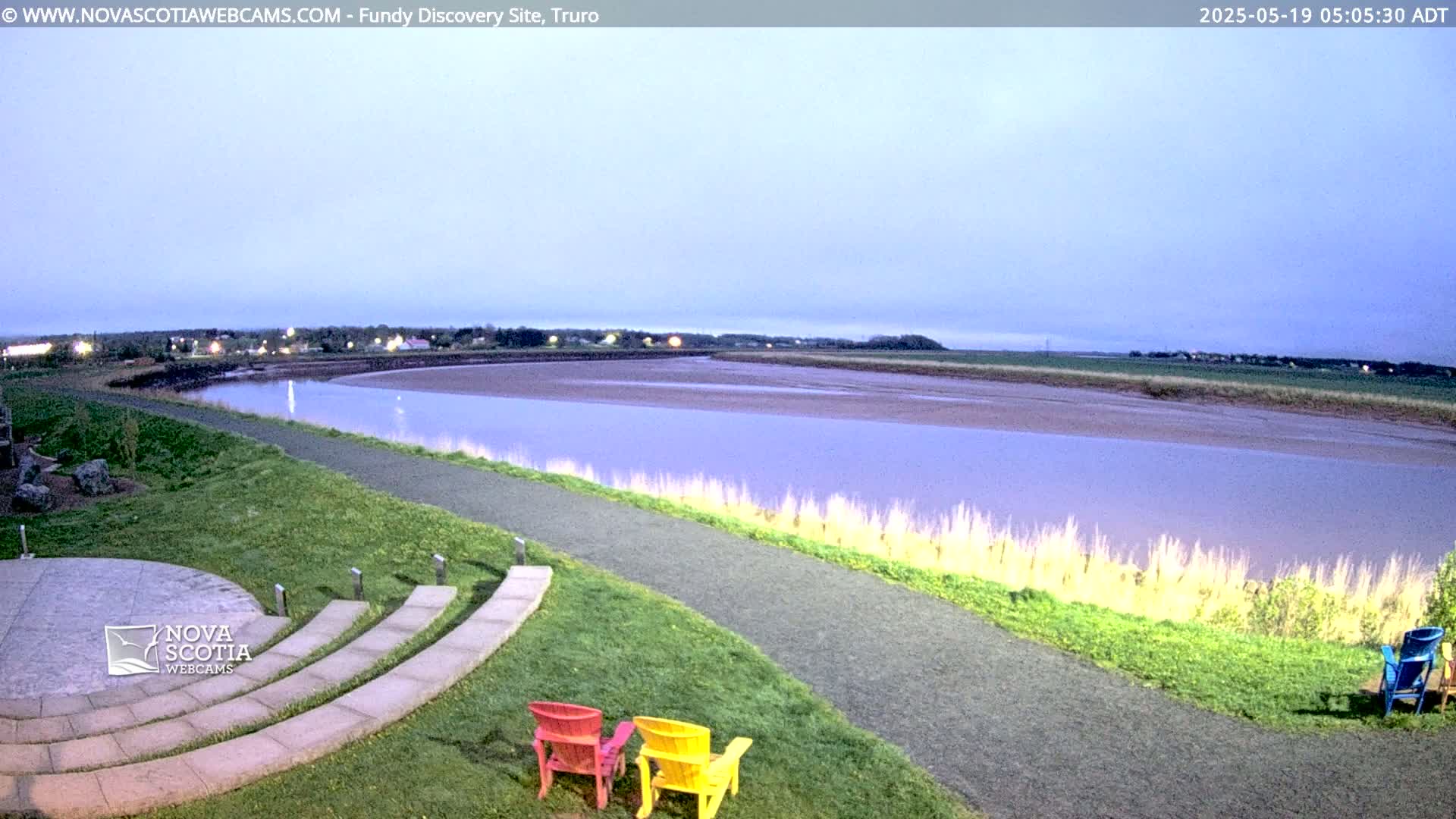 A wide, calm river at low tide is viewed from a grassy area with an amphitheater and several colorful chairs under a twilight sky.