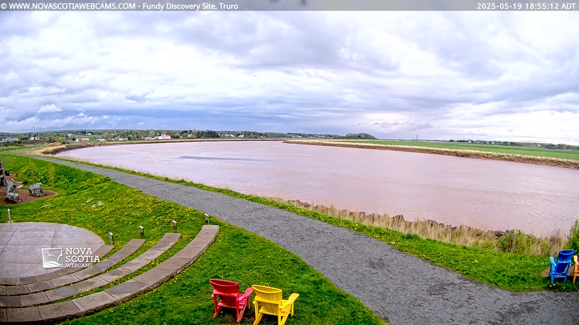 A wide, pinkish-brown river flows under a partly cloudy sky, with a paved pathway leading to a grassy area containing several colorful chairs and a stone amphitheater.