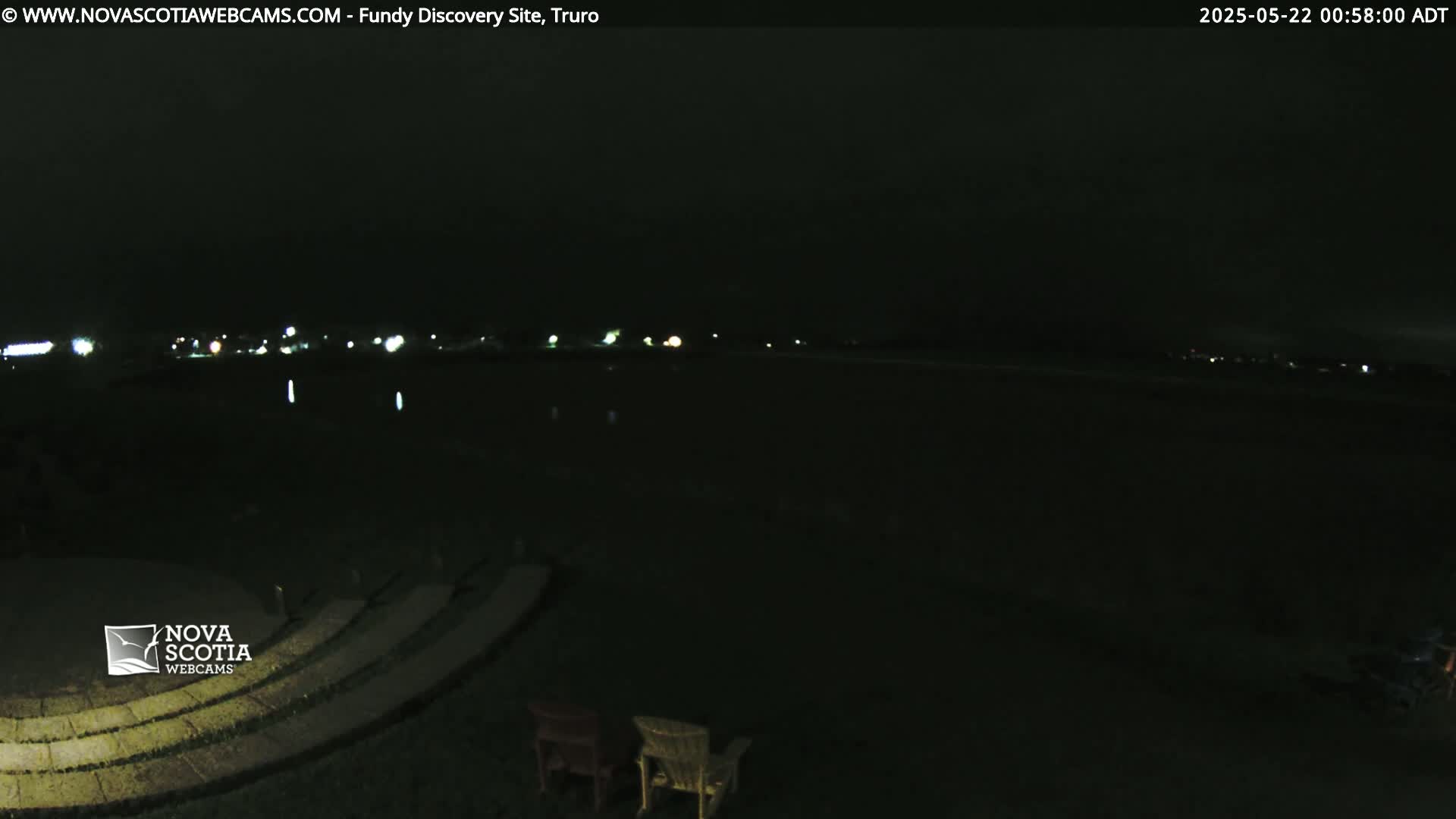 At night, a dark body of water is visible in the distance, with a few faint lights illuminating a shoreline, and two empty chairs sit in the foreground on a stone amphitheater.