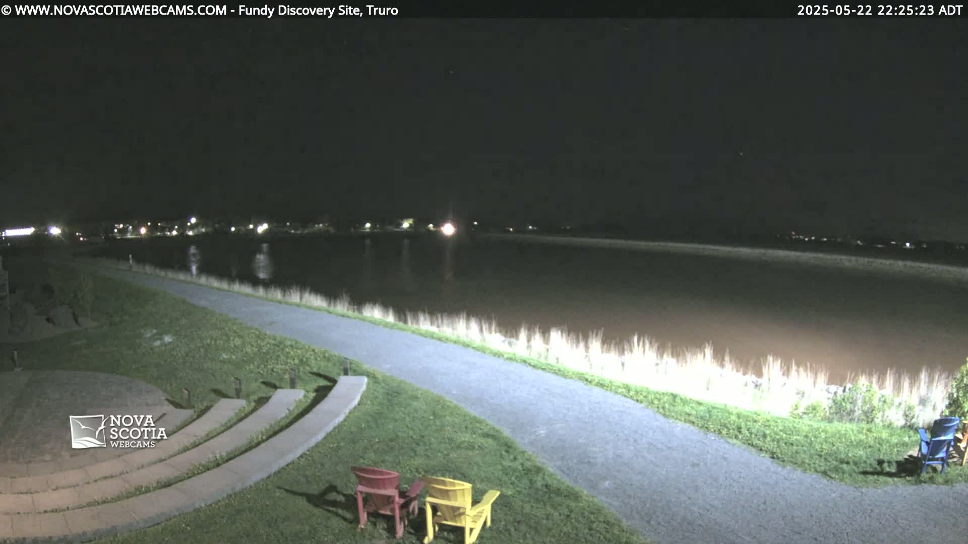 A paved pathway overlooks a calm body of water at night, with distant lights visible across the water and two empty chairs situated on a grassy area near the path.