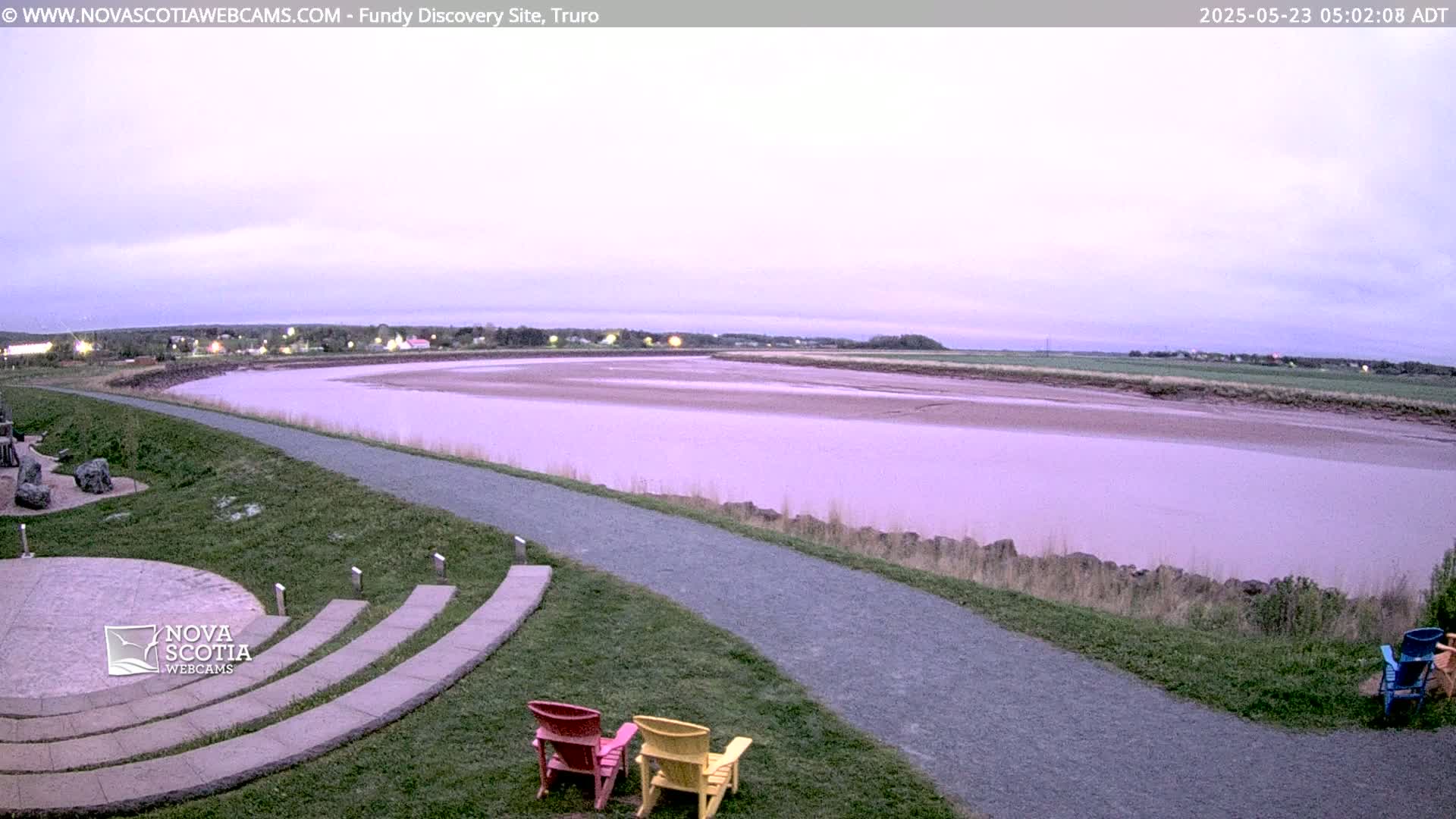 A wide, calm river at low tide stretches out under a pale purple twilight sky, with a paved pathway and a few chairs in the foreground near a grassy area.