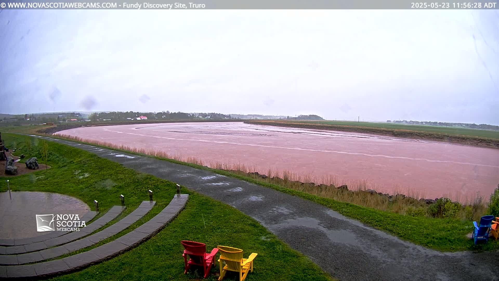 A pink river flows under an overcast sky, beside a paved path and grassy area with several colorful Adirondack chairs.