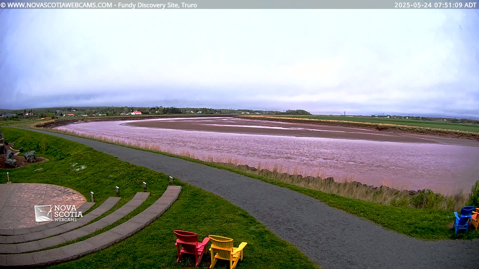 A wide, muddy river flows under an overcast sky, alongside a paved path and grassy area with a few colorful Adirondack chairs.
