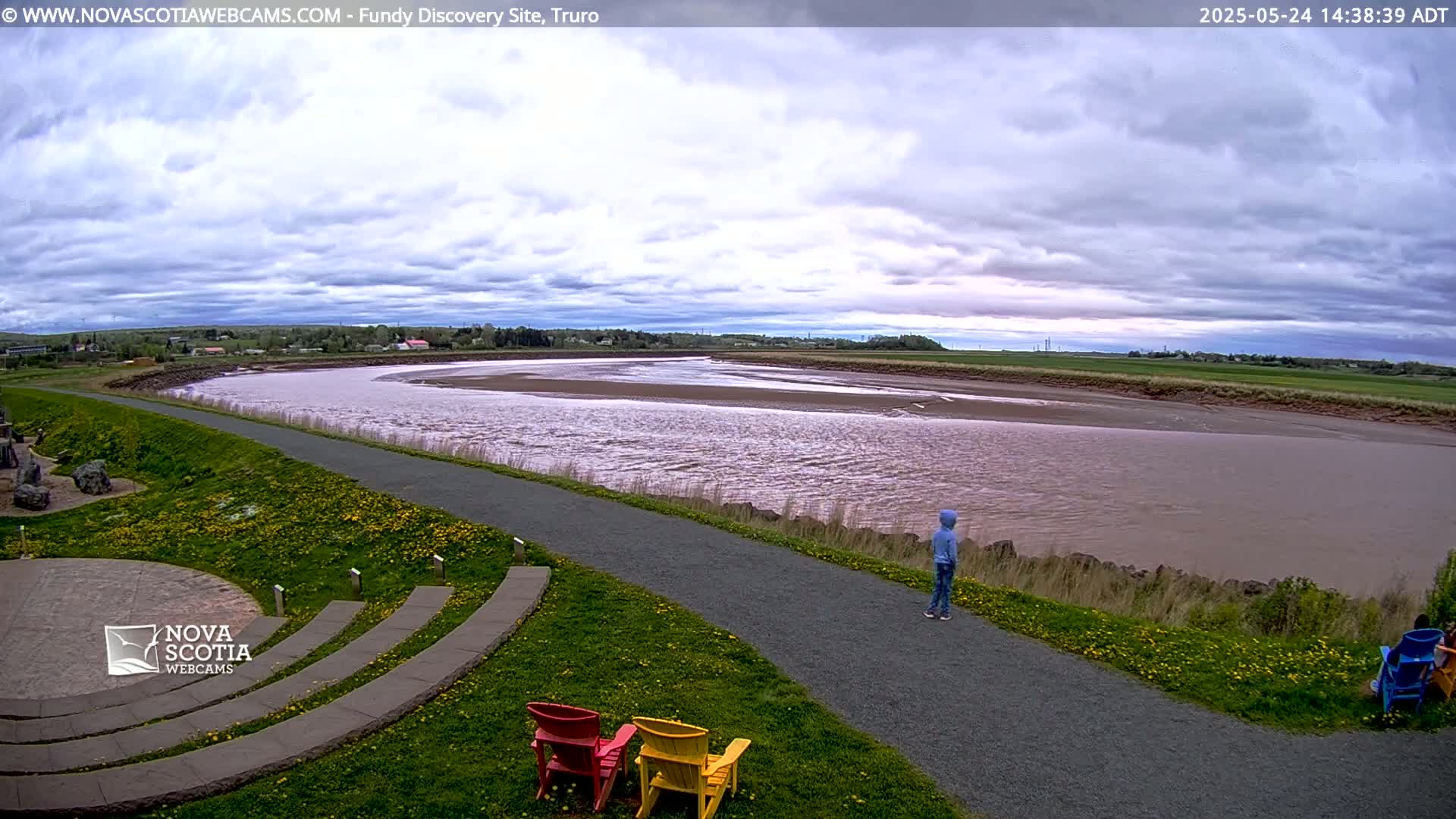 A wide, muddy river flows under a cloudy sky, past a paved walkway with a few people and some empty chairs beside a grassy area.