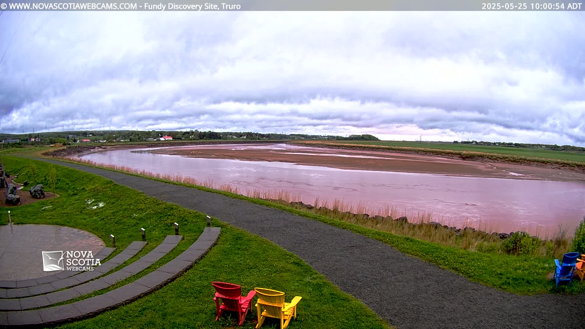 A wide, pinkish-red river flows through a flat landscape under a cloudy sky, with a paved path and colorful Adirondack chairs in the foreground.