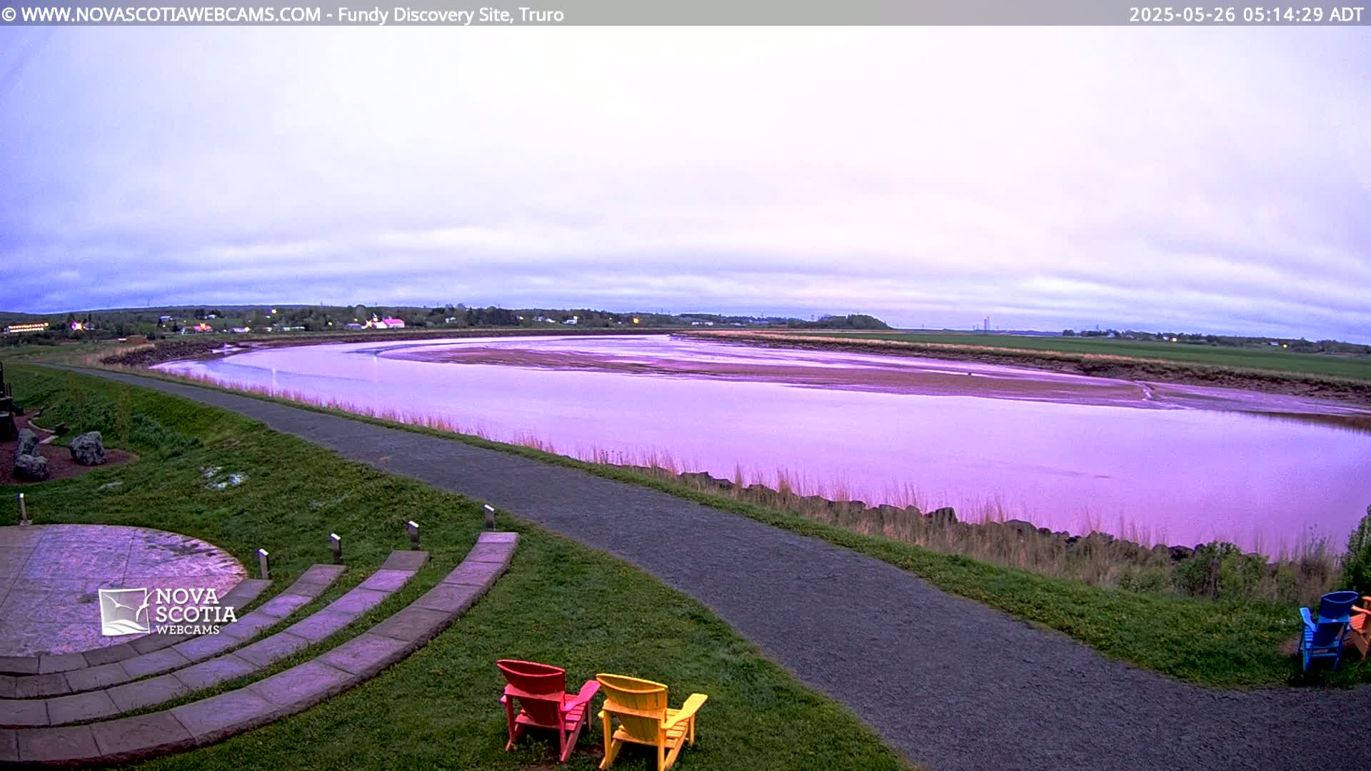 A wide, calm river, appearing purplish under a cloudy, pale sky, is viewed from a grassy area with a paved path and several colorful chairs.