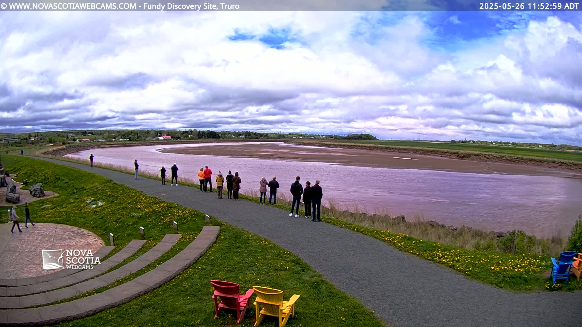 A wide, muddy river under a partly cloudy sky is viewed from a paved walkway where several people stand near colorful Adirondack chairs.