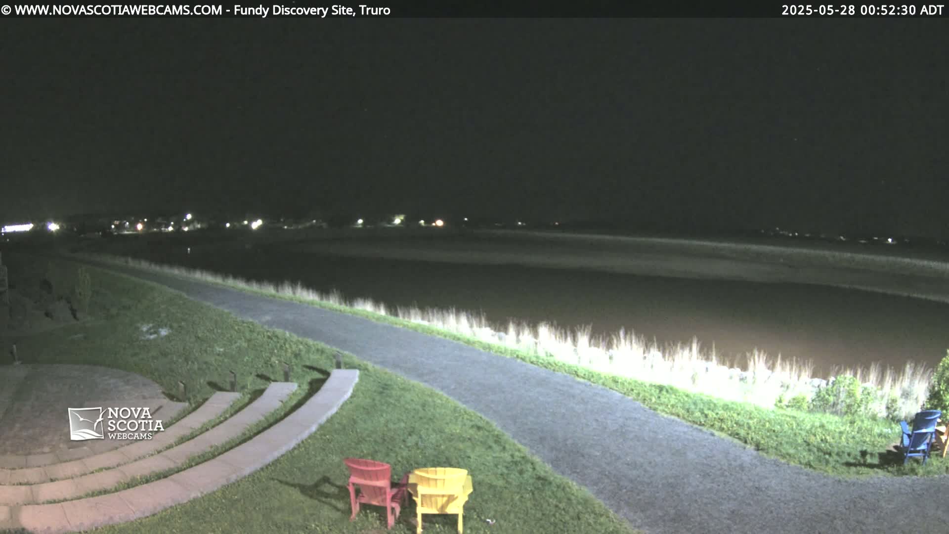 A nighttime view of a paved path curving alongside a calm body of water, with a stone amphitheater and brightly colored chairs in the foreground under a dark sky.