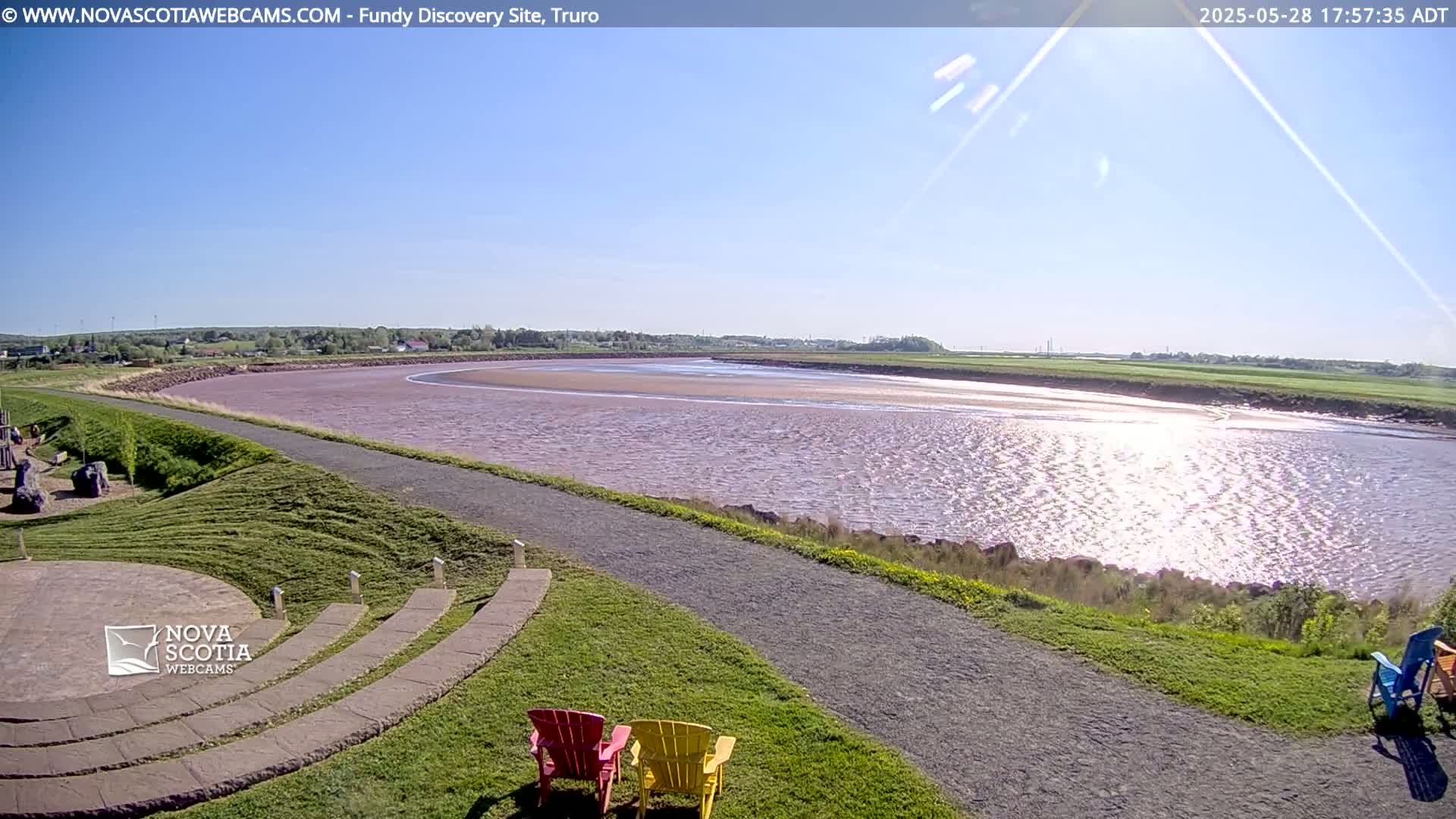 A sunny day reveals a wide, calm river reflecting sunlight, a grassy area with a paved pathway and a few colorful chairs, and a distant, flat landscape under a clear blue sky.