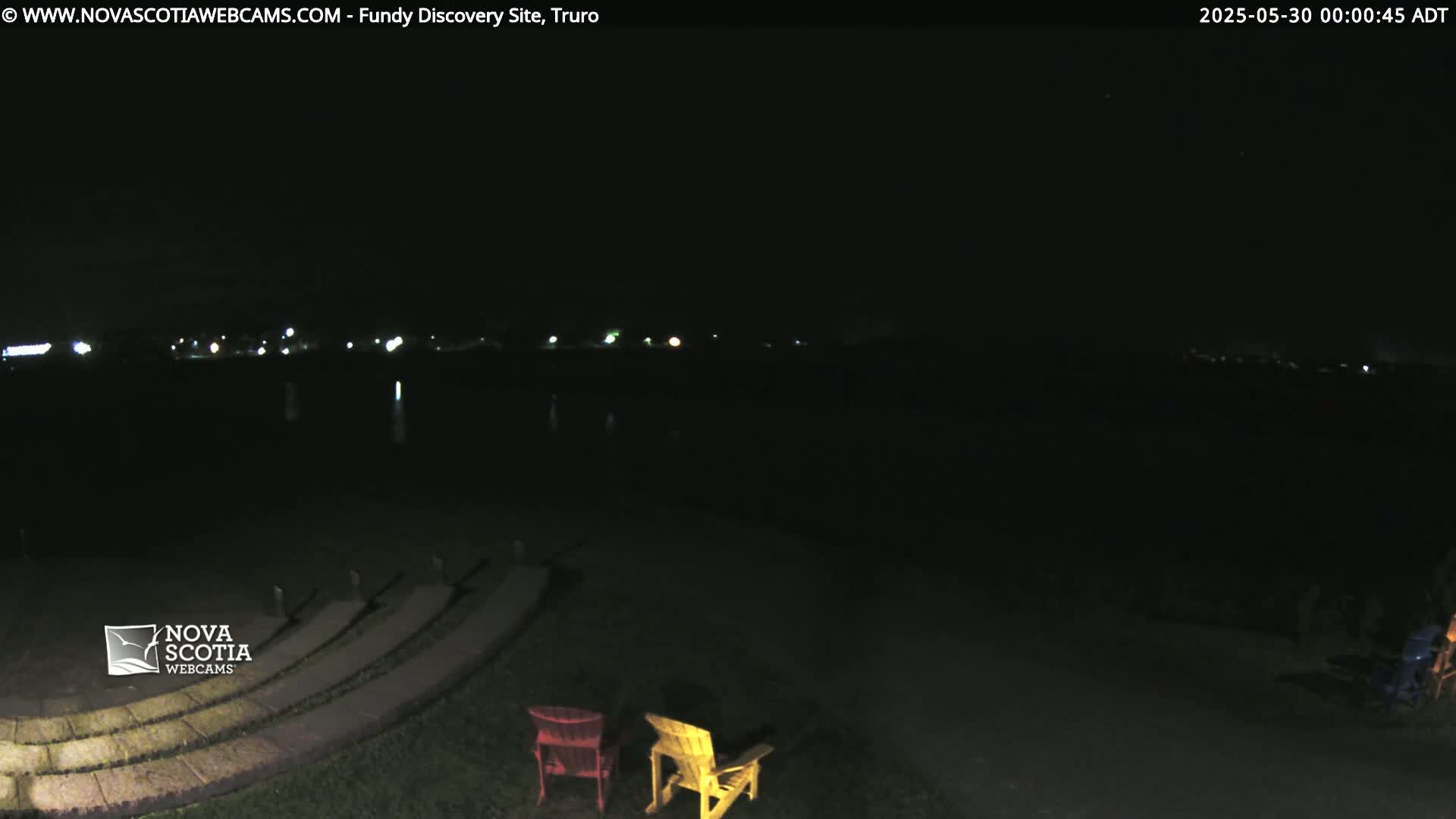 A dark nighttime scene shows a stone amphitheater with two Adirondack chairs and distant lights across a body of water.