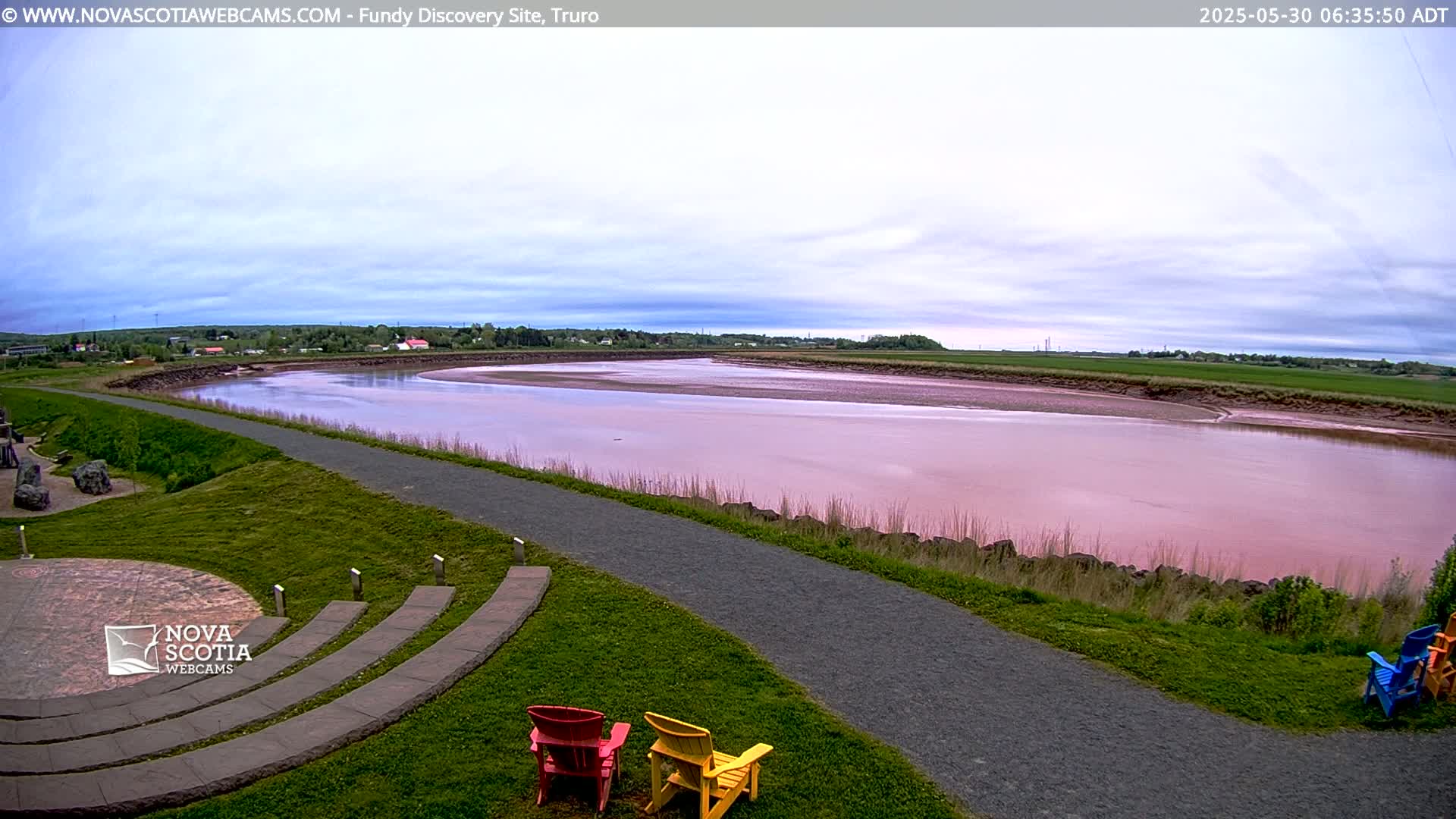 A pink-tinged river flows through a flat landscape under a mostly cloudy sky, with a paved path and a few colorful lawn chairs visible in the foreground.