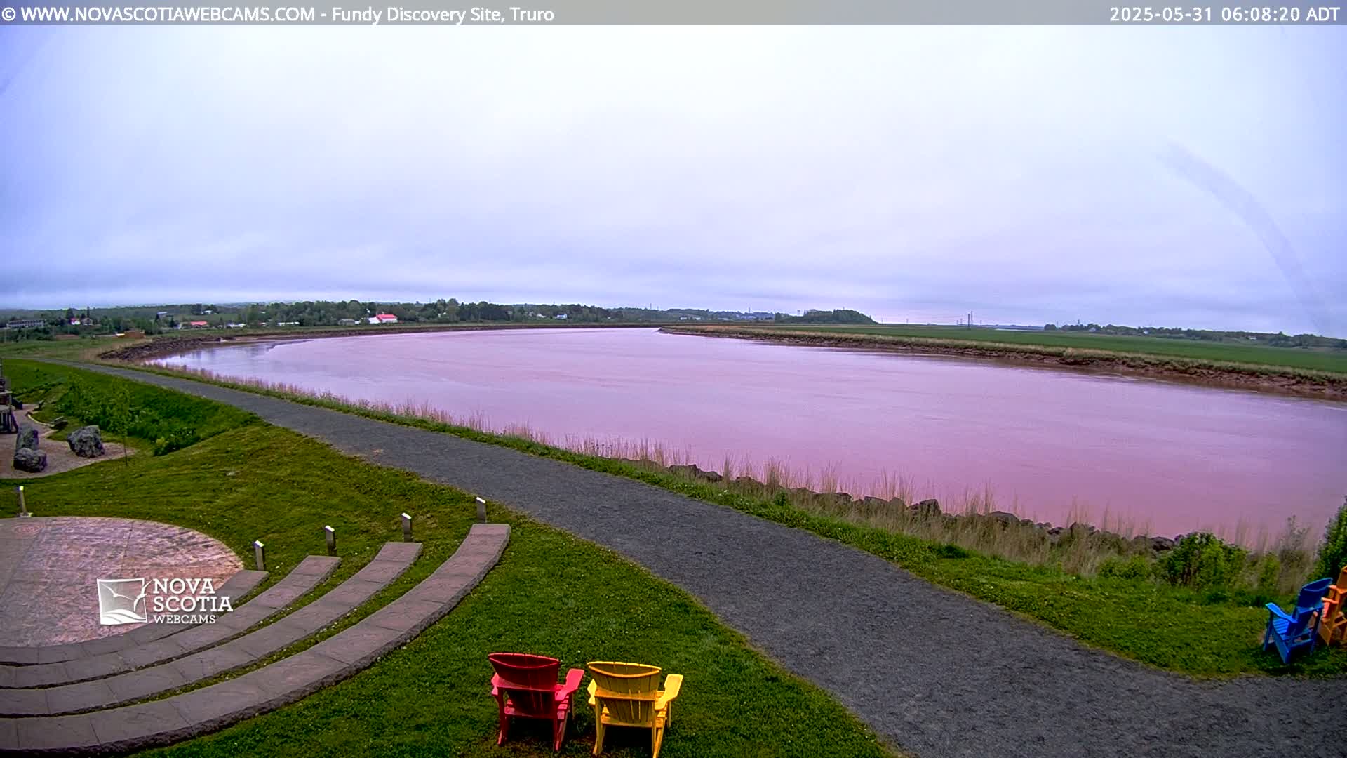 A pink river curves through a grassy landscape under an overcast sky, with a paved walkway and several colorful chairs in the foreground.