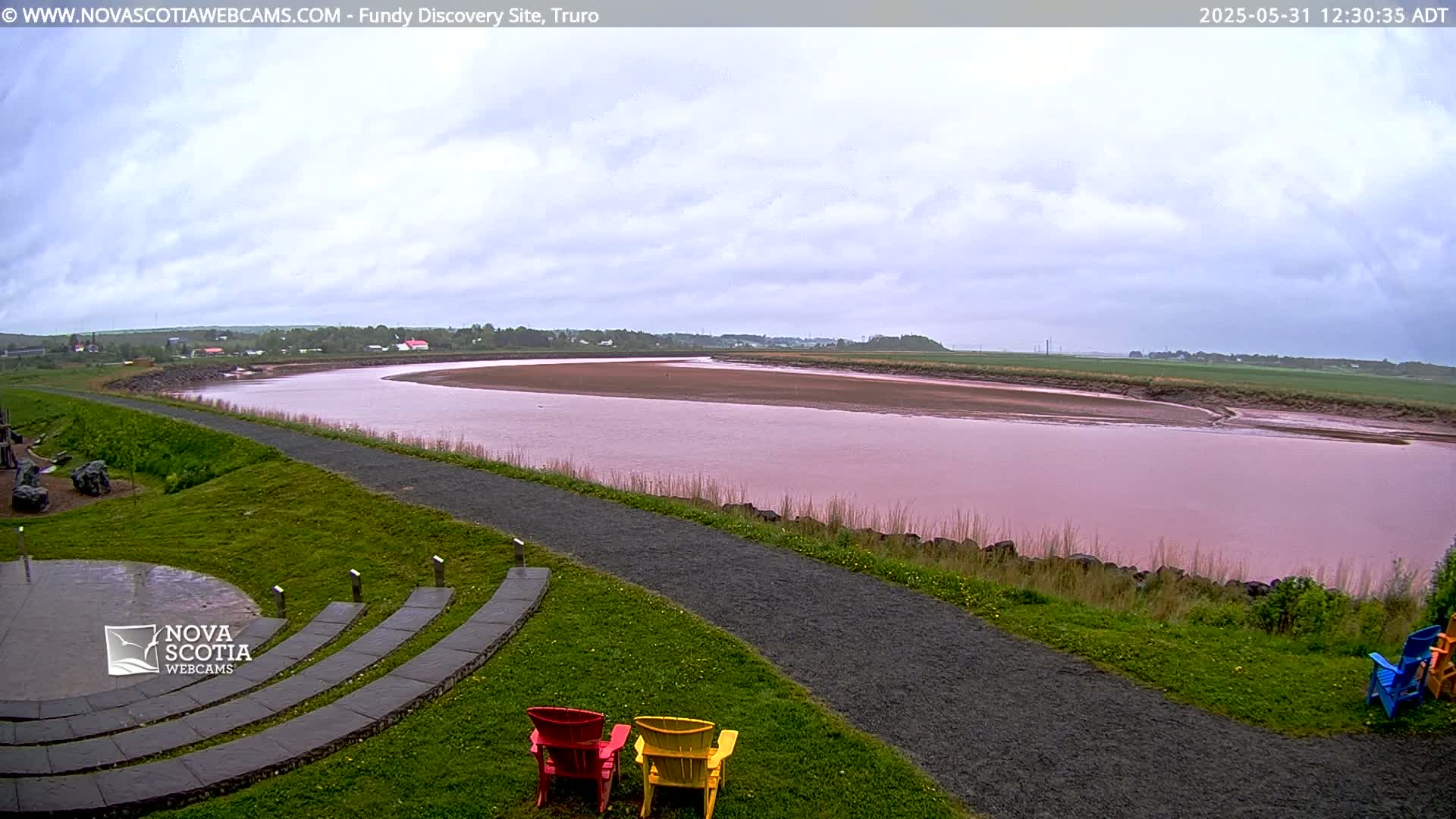 A pink river curves through a green landscape under a cloudy sky, with a stone amphitheater and colorful Adirondack chairs in the foreground.