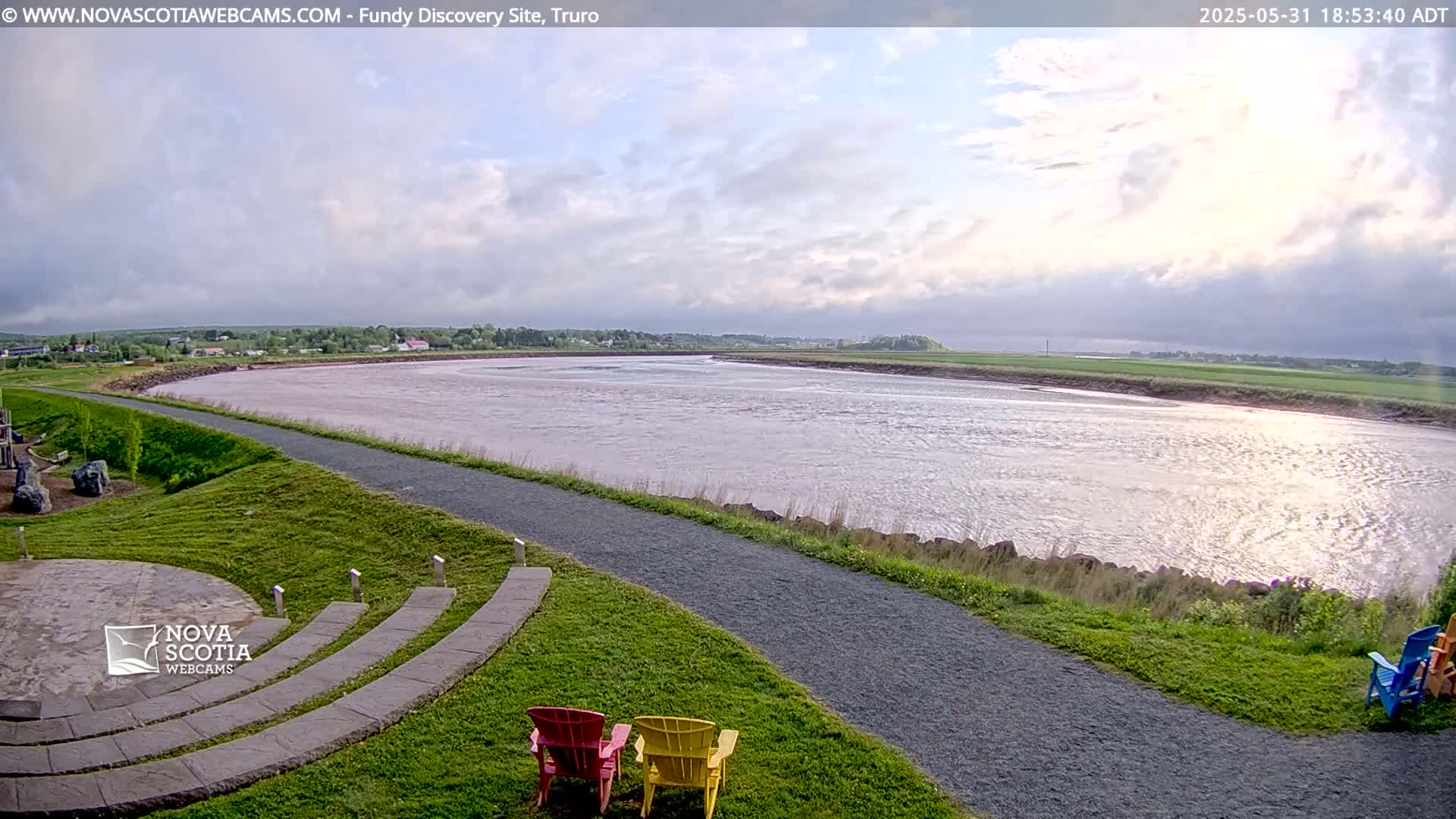 A wide, muddy river flows under a partly cloudy sky, with a paved path and a few colorful lawn chairs in the foreground.