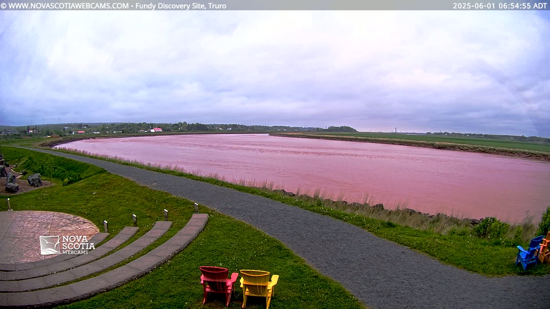 A pink river flows under an overcast sky, alongside a paved pathway and grassy area with several colorful Adirondack chairs.