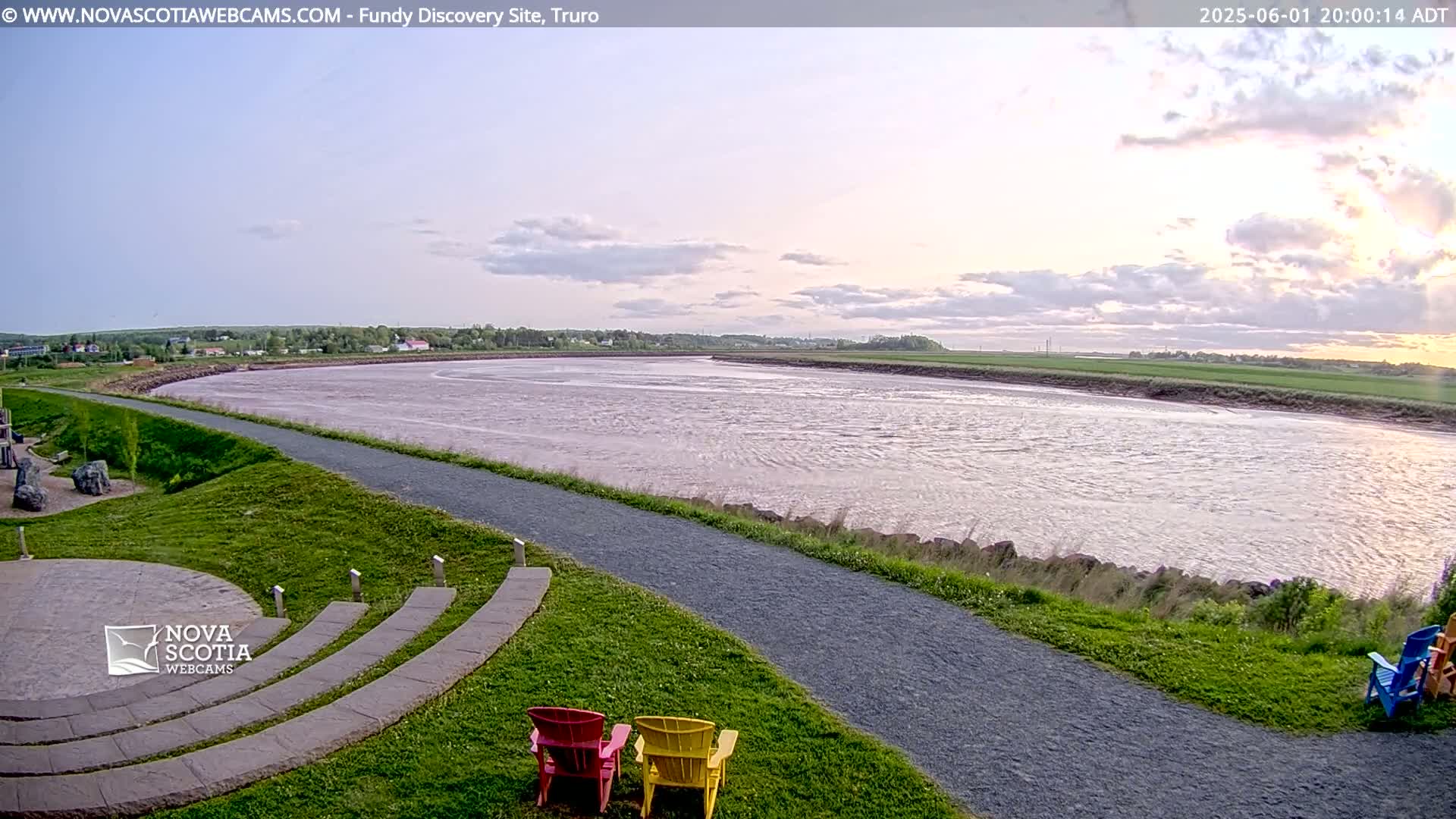 A wide, calm river flows under a partly cloudy, sunset sky past a grassy area with a paved walkway and several colorful chairs.