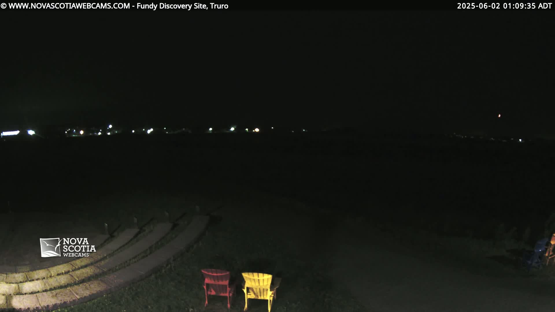 At night, two empty Adirondack chairs sit in front of an empty stone amphitheater, overlooking a distant line of lights under a dark, clear sky.