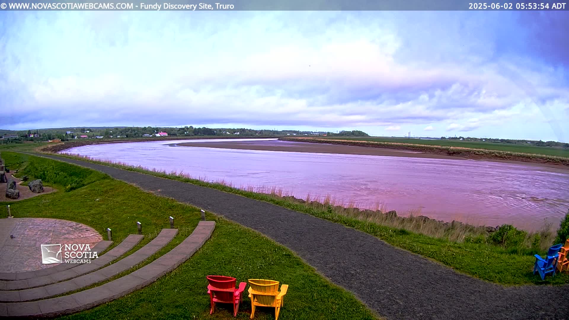A wide, pinkish-brown river curves gently through a green landscape under a partly cloudy sky.