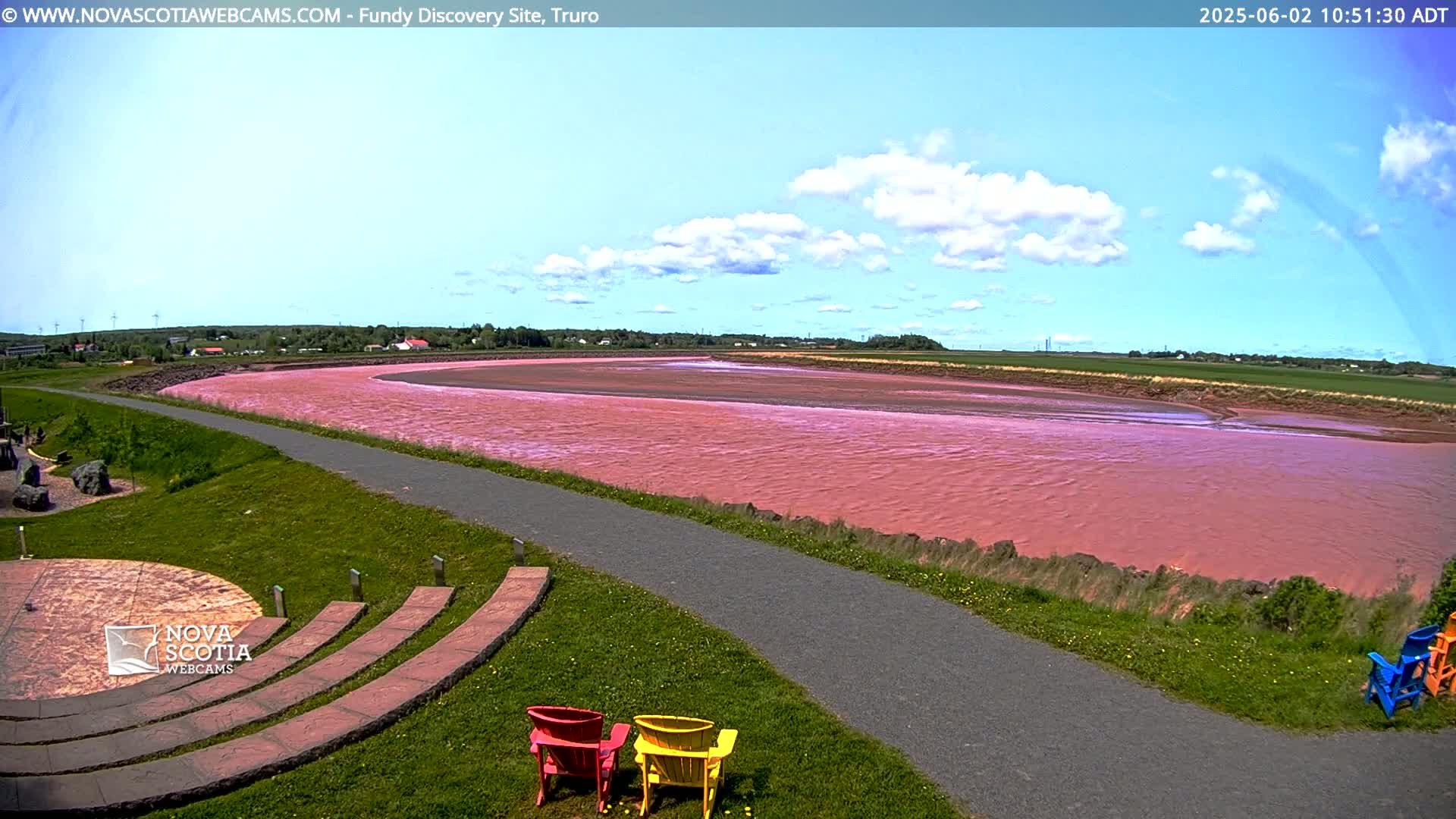 A pink river flows through a green landscape under a partly cloudy blue sky, with a paved path and colorful chairs visible in the foreground.