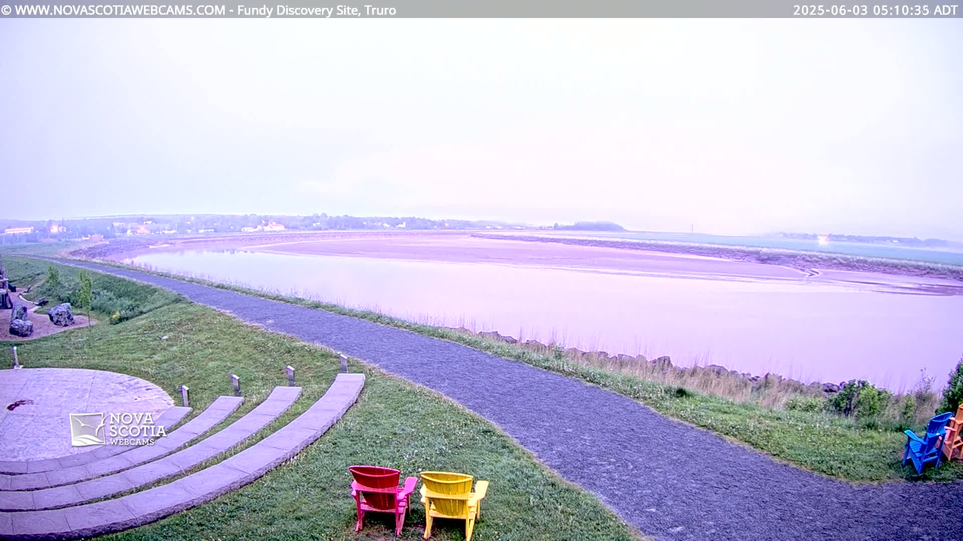A wide, calm river under a hazy, pale sky is viewed from a grassy area with a paved pathway, stone seating, and a few colorful chairs.