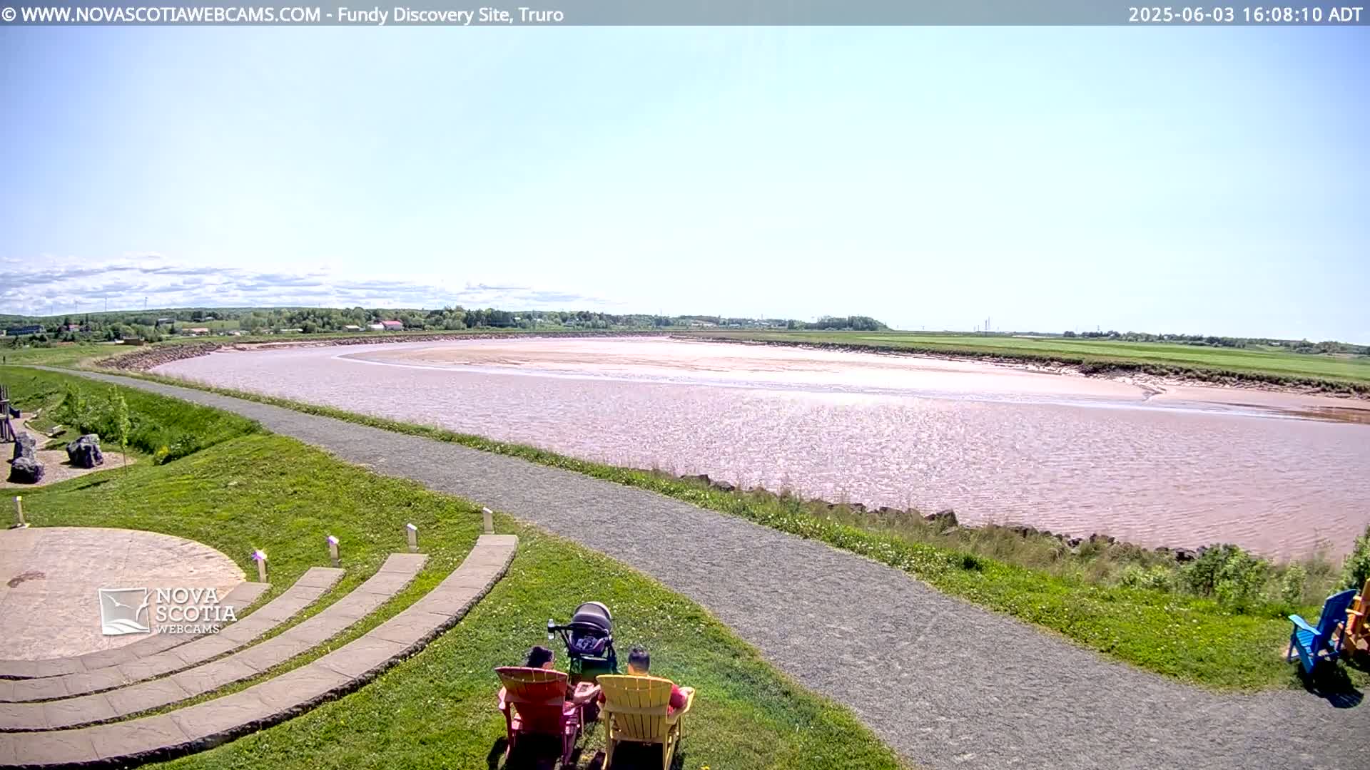 A family sits in chairs overlooking a wide, calm, pinkish-brown river under a mostly sunny sky.