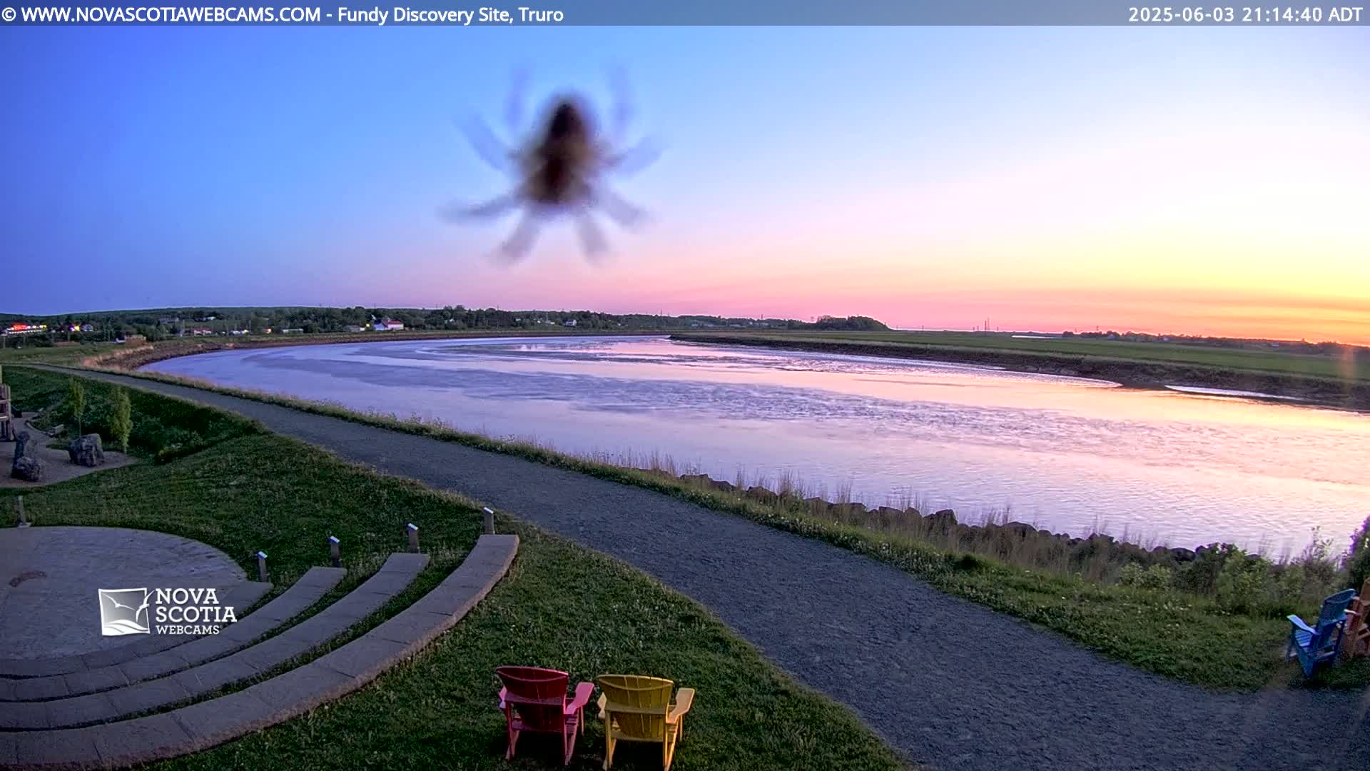 A blurred spider is in the foreground of a tranquil river scene at sunset, with a paved walking path and Adirondack chairs visible in the lower right.