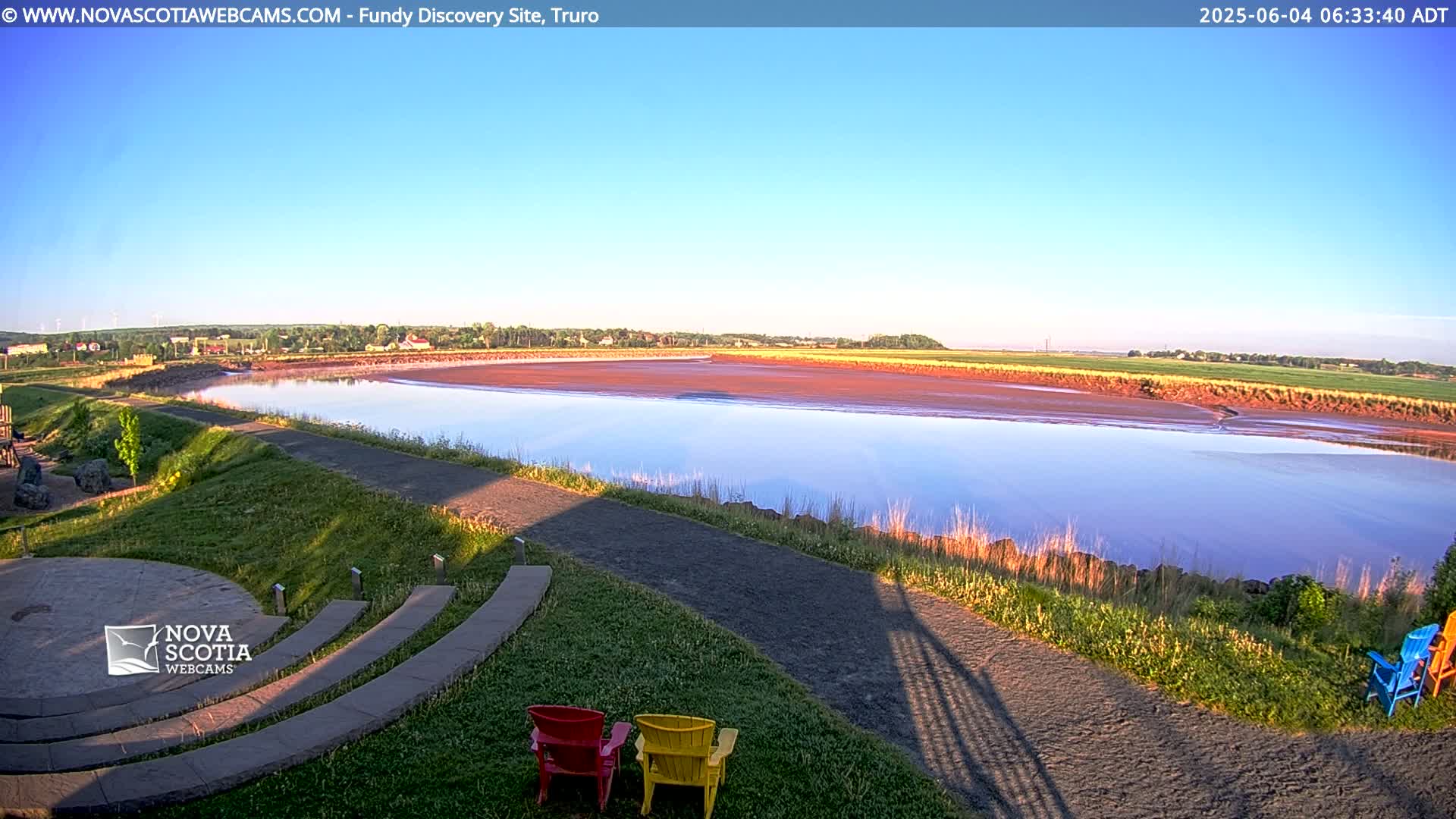 A calm river with a reddish-brown mudflat stretches toward a distant town under a clear blue sky.