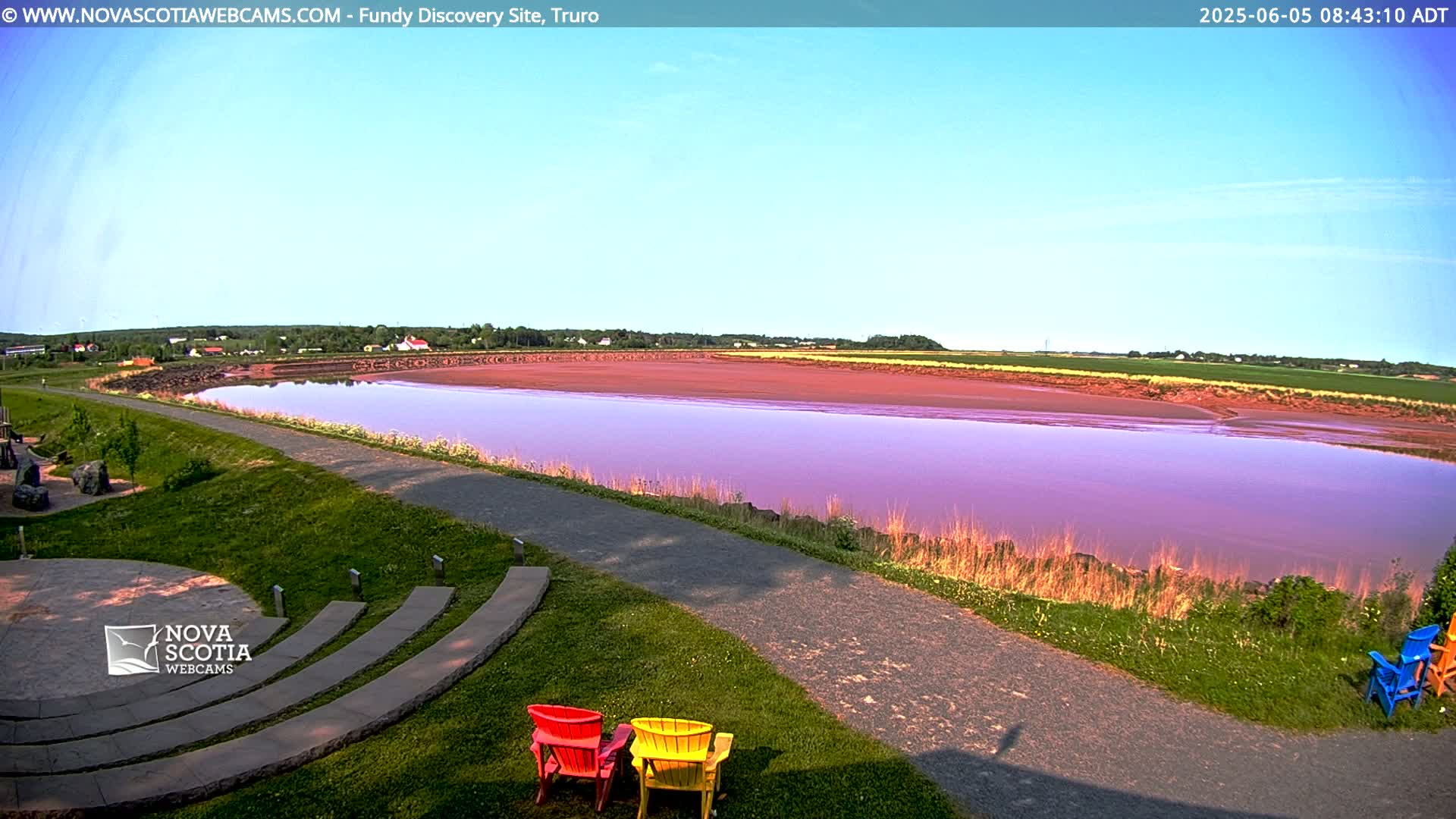 A wide, calm body of purplish-pink water stretches across a flat landscape under a clear, light-blue sky, with grassy areas and a paved pathway in the foreground, and a few colorful chairs near a stone amphitheater.