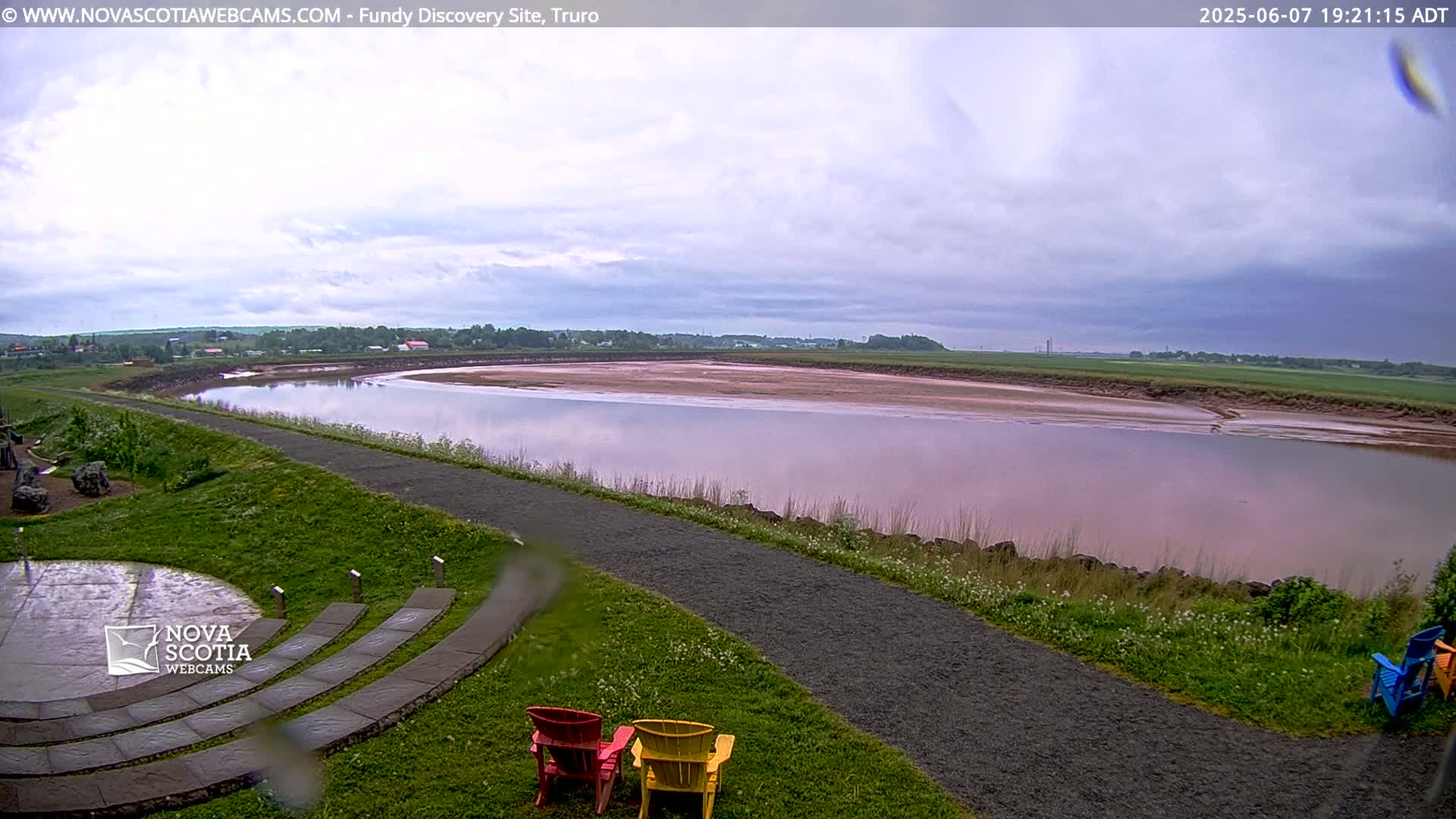 A wide, calm river with pinkish-red mudflats is visible under a cloudy sky, with a paved path and colorful lawn chairs in the foreground.
