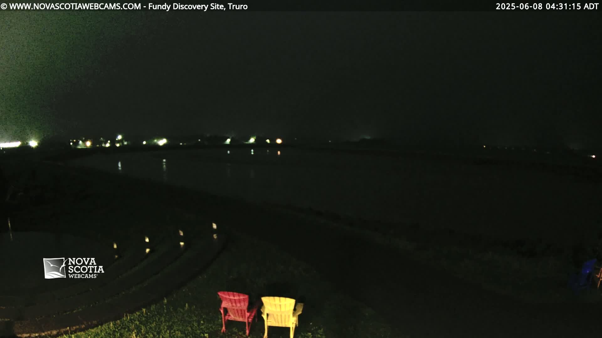 At night, two brightly colored chairs sit on a grassy area overlooking a dark body of water with faintly visible lights across it under a dark, possibly overcast sky.