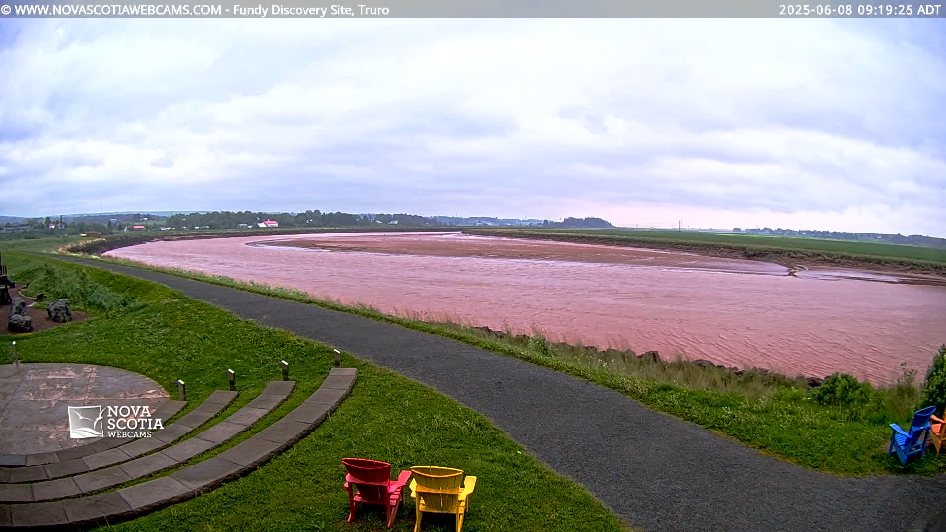 A pinkish-red river flows under an overcast sky, beside a paved path and grassy area with several colorful chairs.