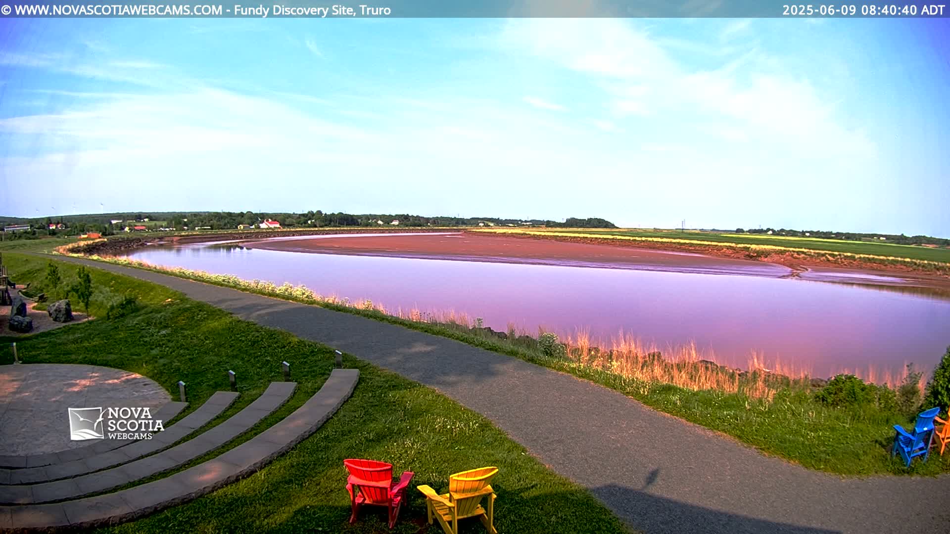 A calm, pinkish-purple river flows through a grassy landscape under a mostly clear, light-blue sky, with three brightly colored chairs visible near a paved path.