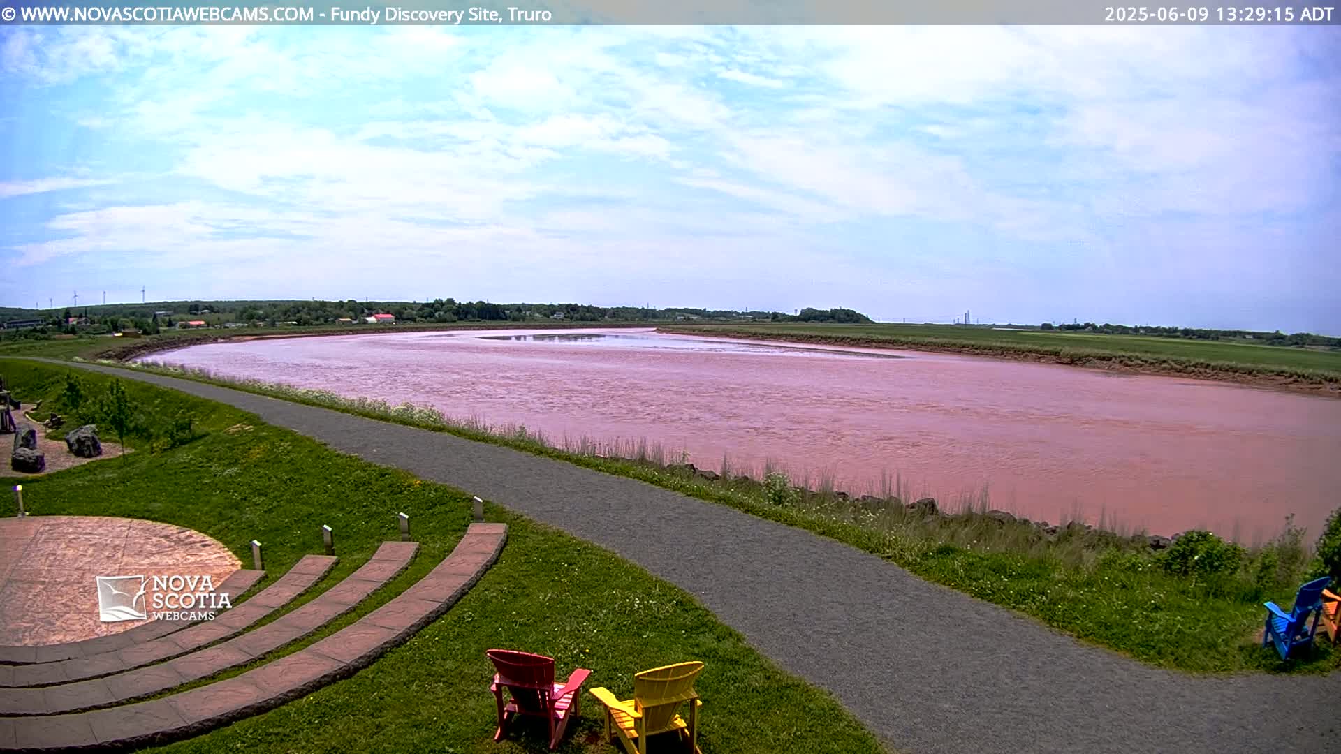A wide, pink river flows through a grassy landscape under a partly cloudy sky, with a paved path and several colorful chairs situated near a stone amphitheater in the foreground.