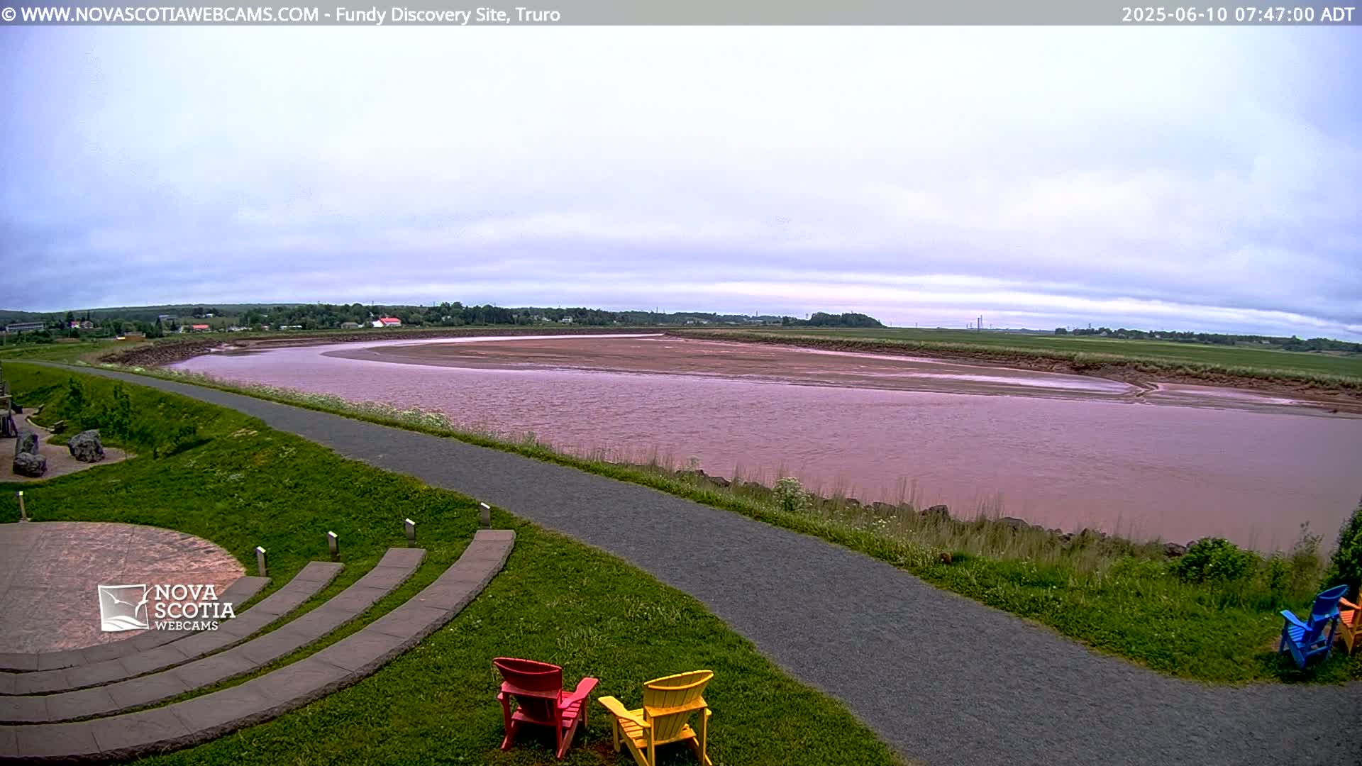 A wide, pinkish-red river curves gently through a grassy landscape under a mostly cloudy sky.