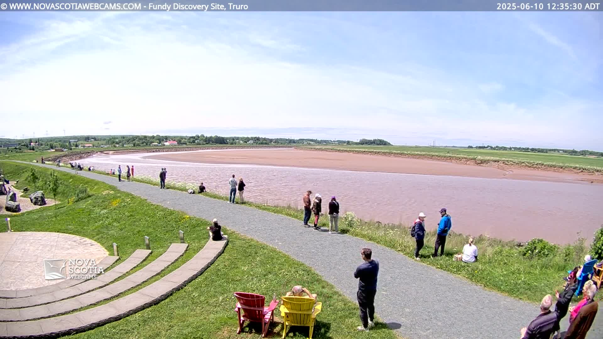 A wide, pink-tinged river flows under a mostly sunny sky, with people gathered on a paved path overlooking the water and enjoying the scenery.