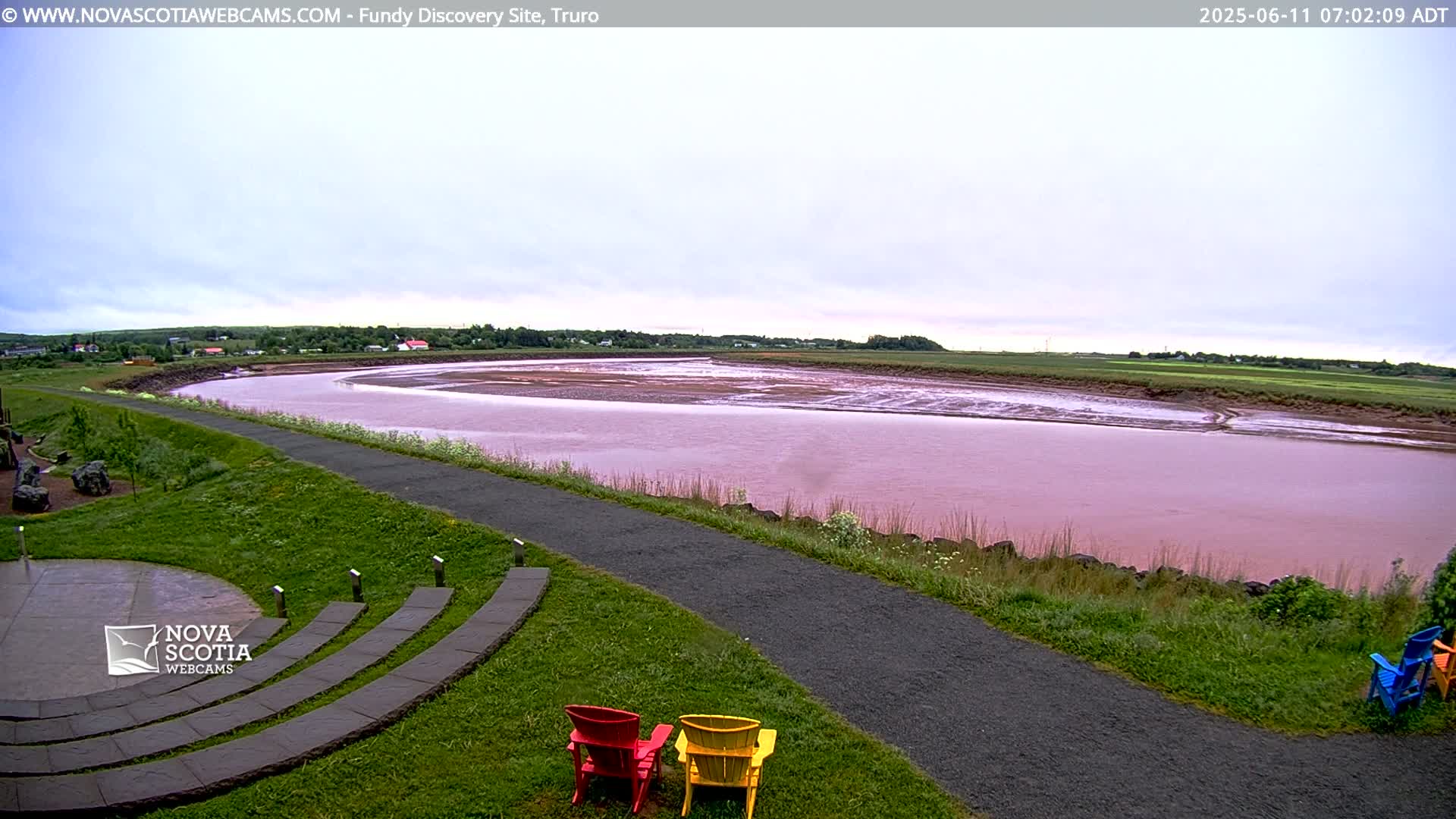 A wide, pinkish-red river curves through a flat landscape under a cloudy sky, with a paved walkway and colorful Adirondack chairs in the foreground.