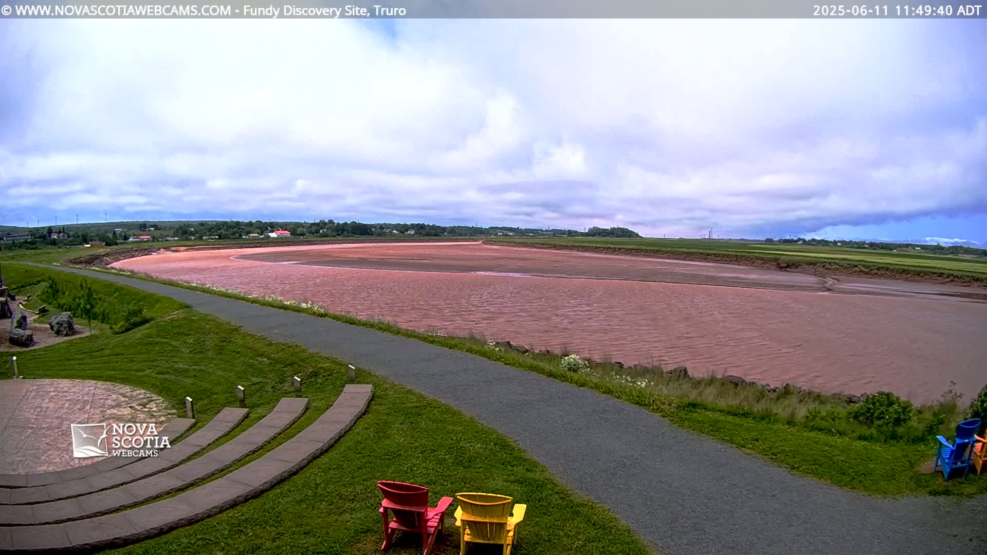 A pinkish-red river flows through a flat landscape under a partly cloudy sky, with a paved path and colorful Adirondack chairs visible in the foreground.