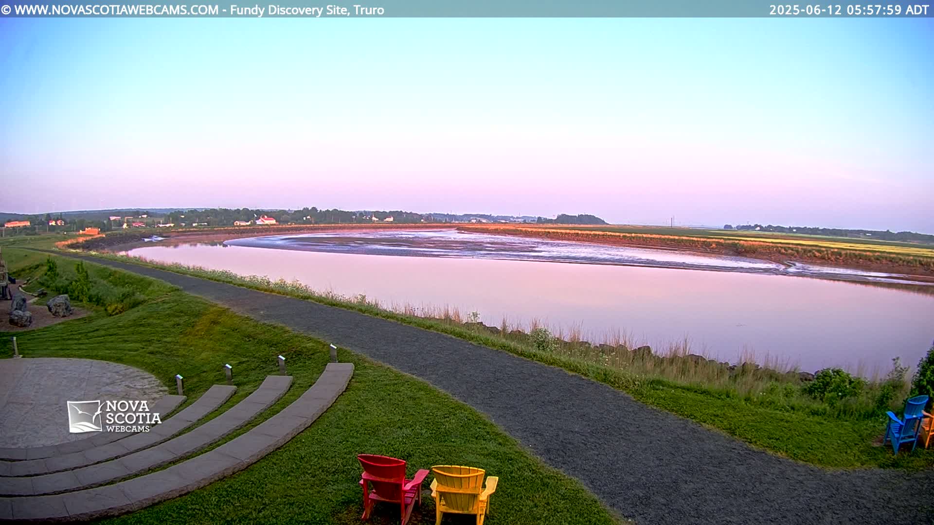 A pink-hued sunrise illuminates a calm river winding through a grassy landscape, with a paved pathway and colorful lawn chairs nearby.