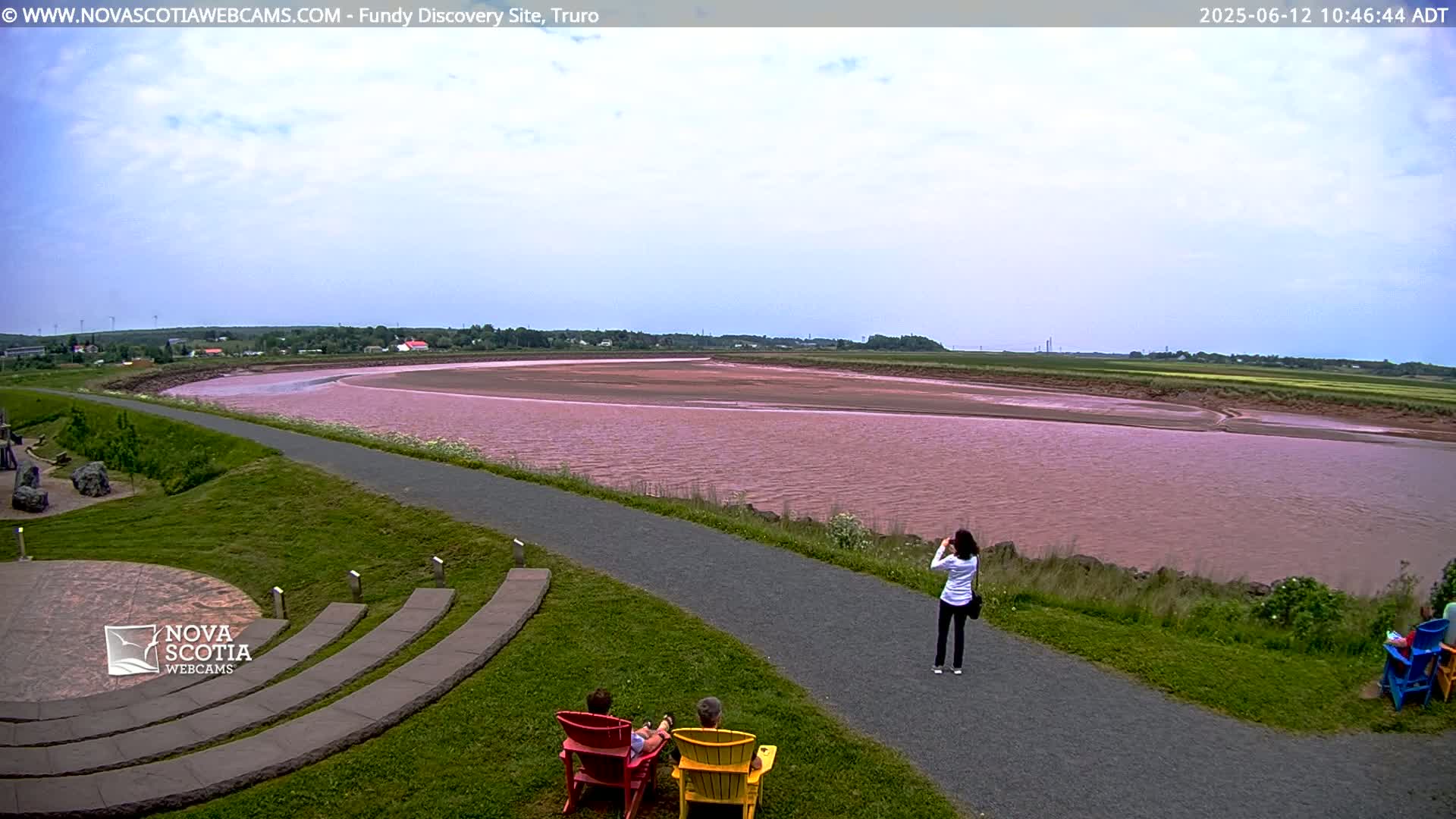 A pinkish-red river flows through a flat landscape under a partly cloudy sky, with people sitting on lawn chairs and walking along a paved path beside it.