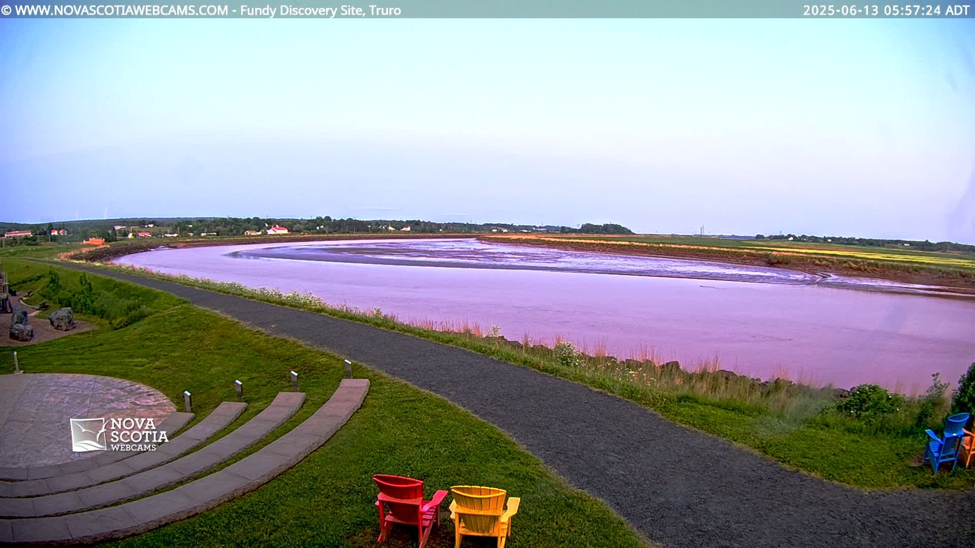 A wide, pinkish river curves through a grassy landscape under a clear, light-blue sky, with a paved pathway and several colorful Adirondack chairs visible in the foreground.