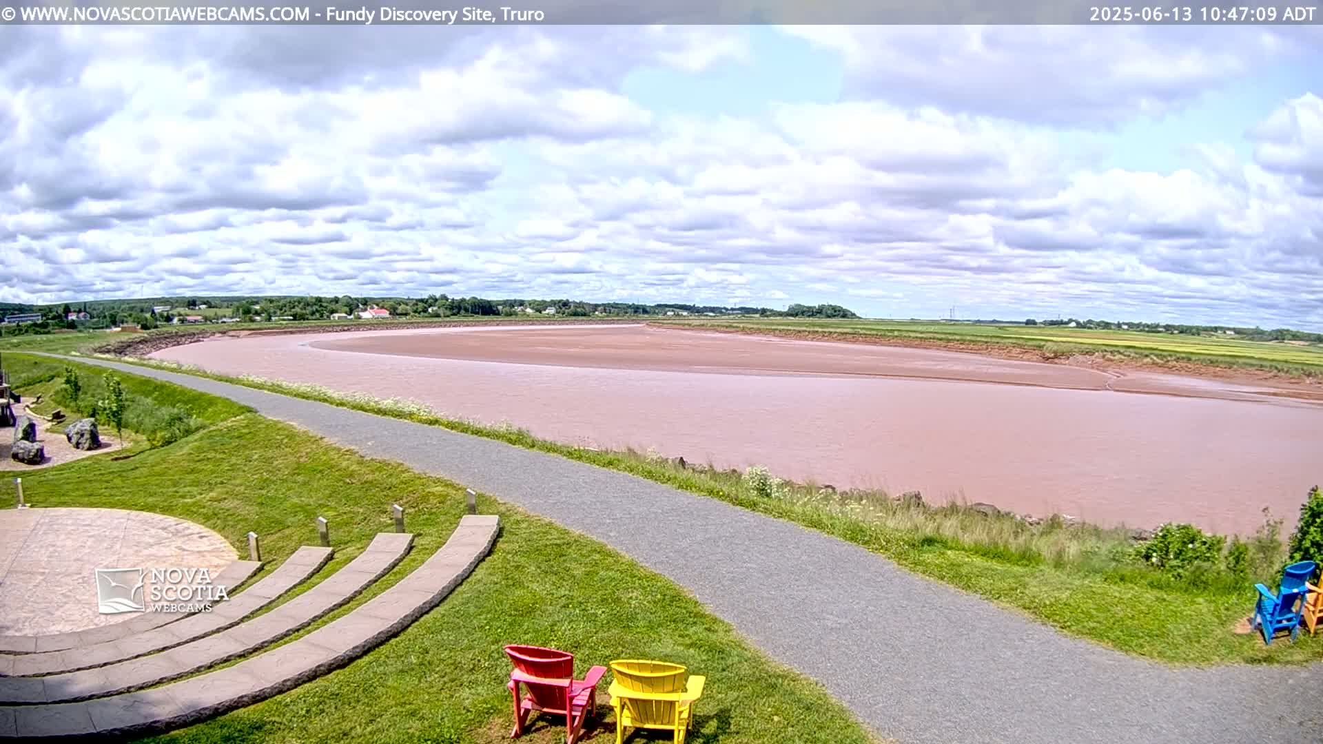 A wide, pink-hued river flows through a flat landscape under a partly cloudy sky, with a paved pathway and amphitheater visible in the foreground.
