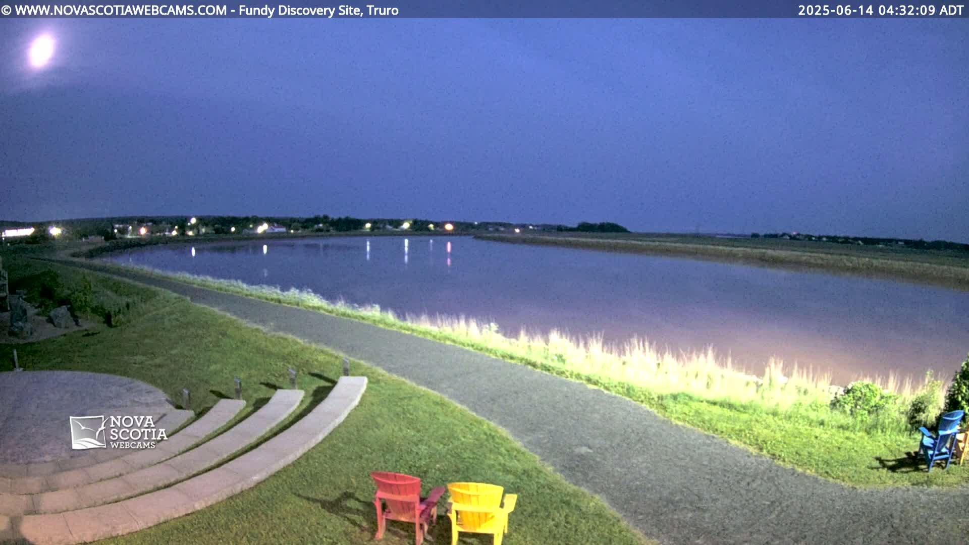 A calm body of water is visible at dusk, next to a paved pathway and amphitheater-style seating, with some colorful chairs placed nearby.