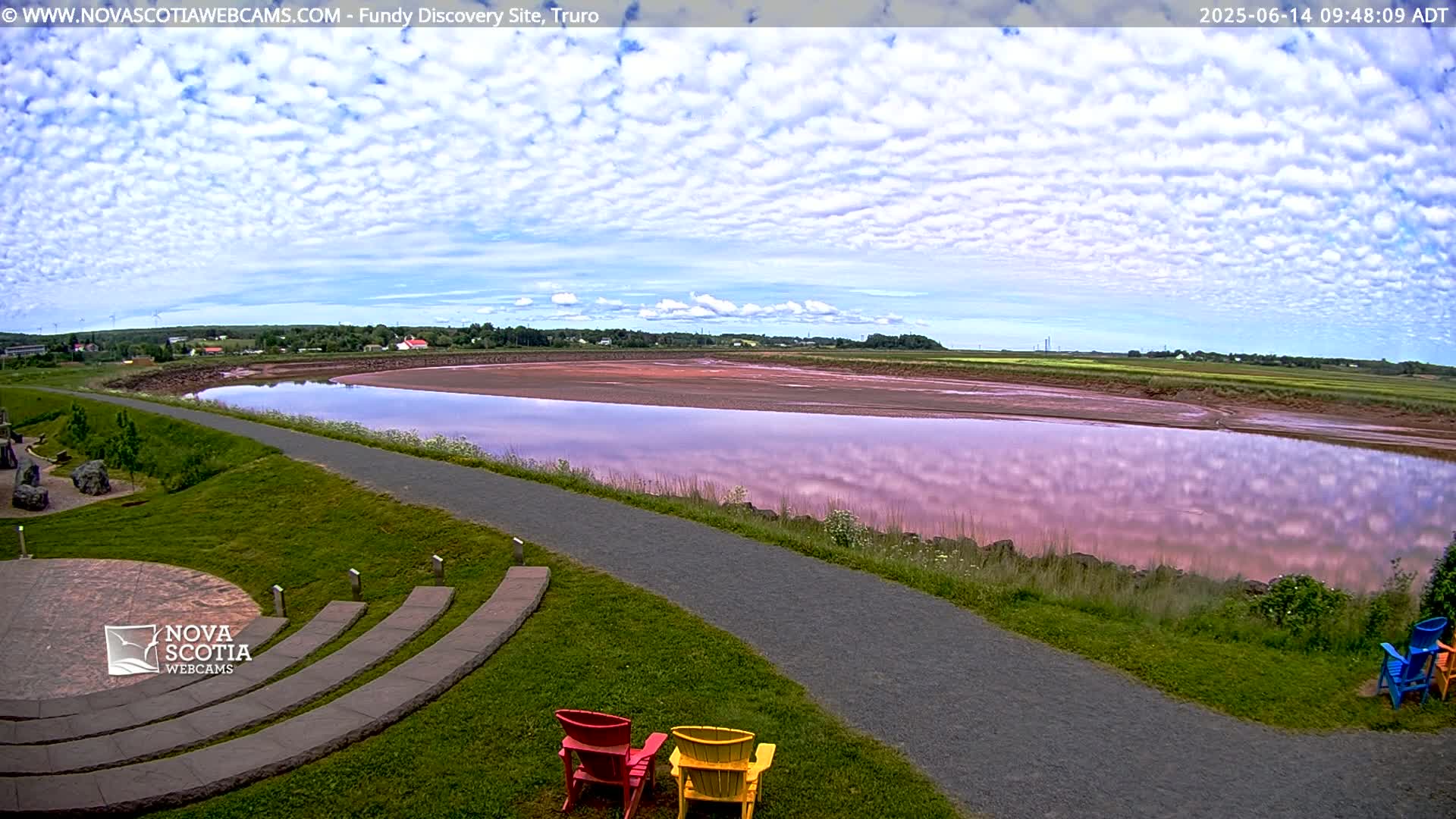 A calm body of pink-tinged water reflects a mostly cloudy sky, alongside a paved pathway and grassy area with several colorful chairs.