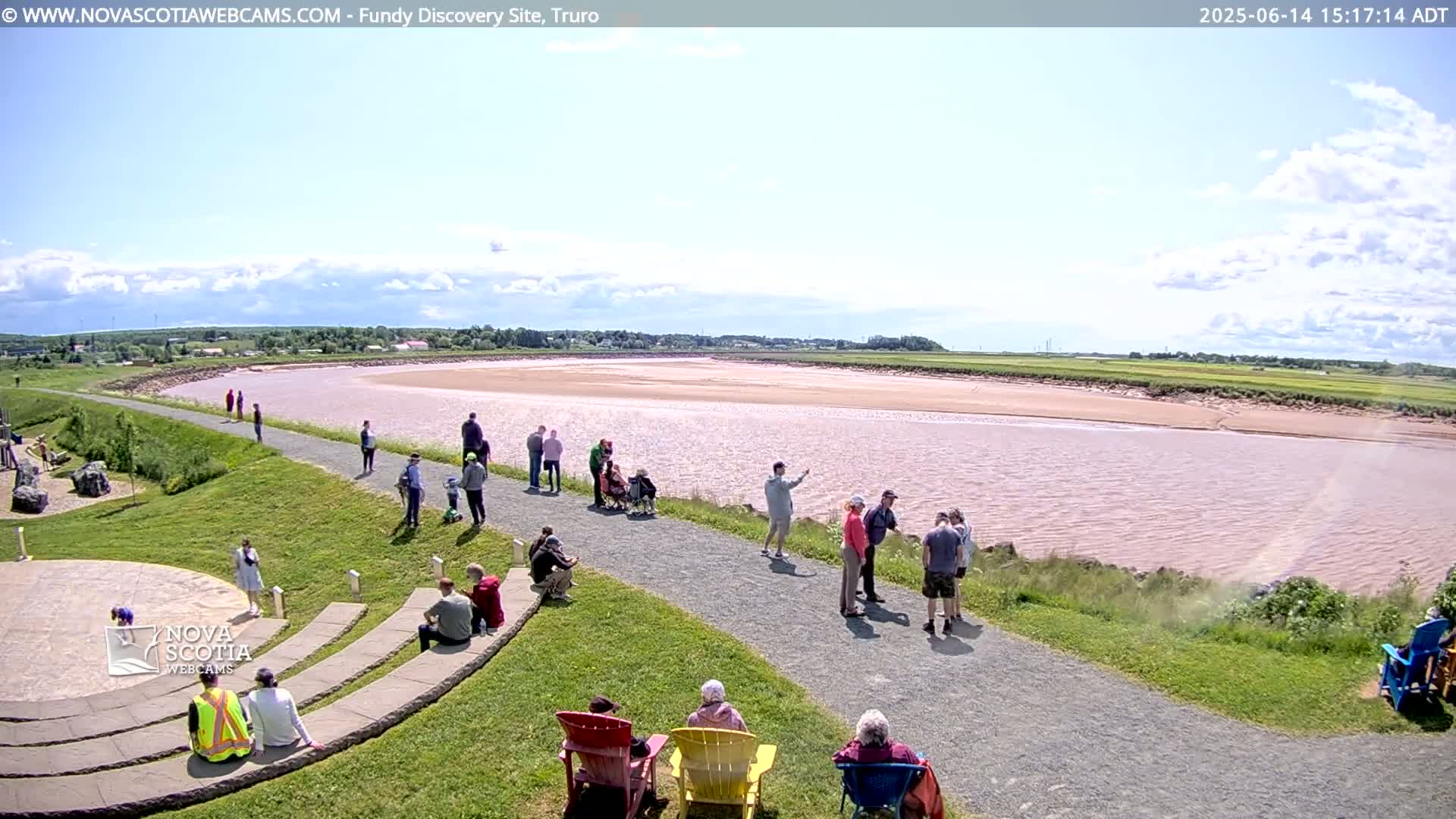 A wide, pink-tinged river stretches out under a partly cloudy sky, with many people gathered on a grassy area and pathway overlooking the water.