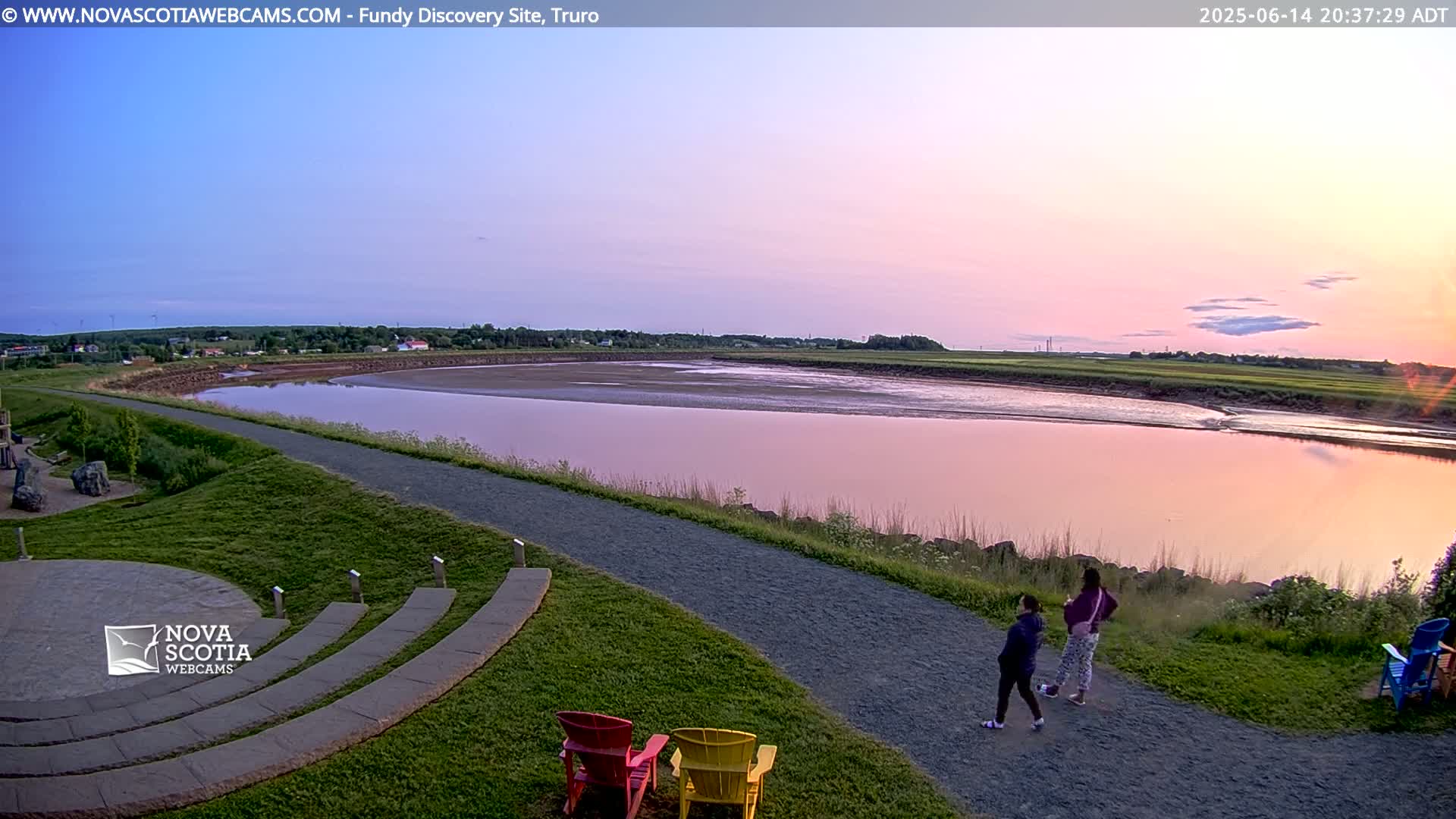 A wide, pink-tinged river stretches out under a partly cloudy sky, with many people gathered on a grassy area and pathway overlooking the water.