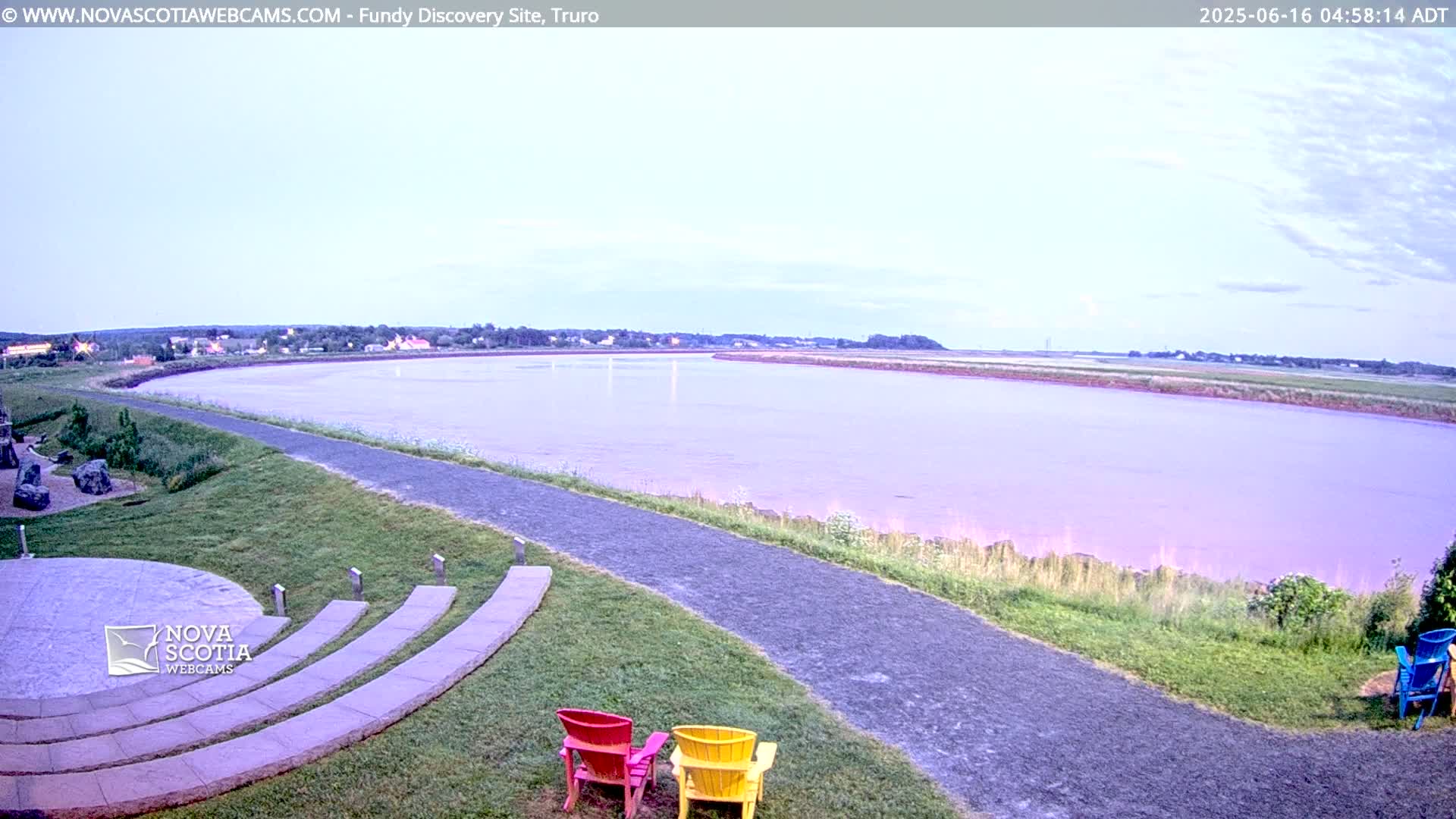 A wide, calm river with a pinkish hue stretches towards a distant town under a light-blue, mostly clear sky, a paved path curves alongside the riverbank towards a stone amphitheater with colorful Adirondack chairs.
